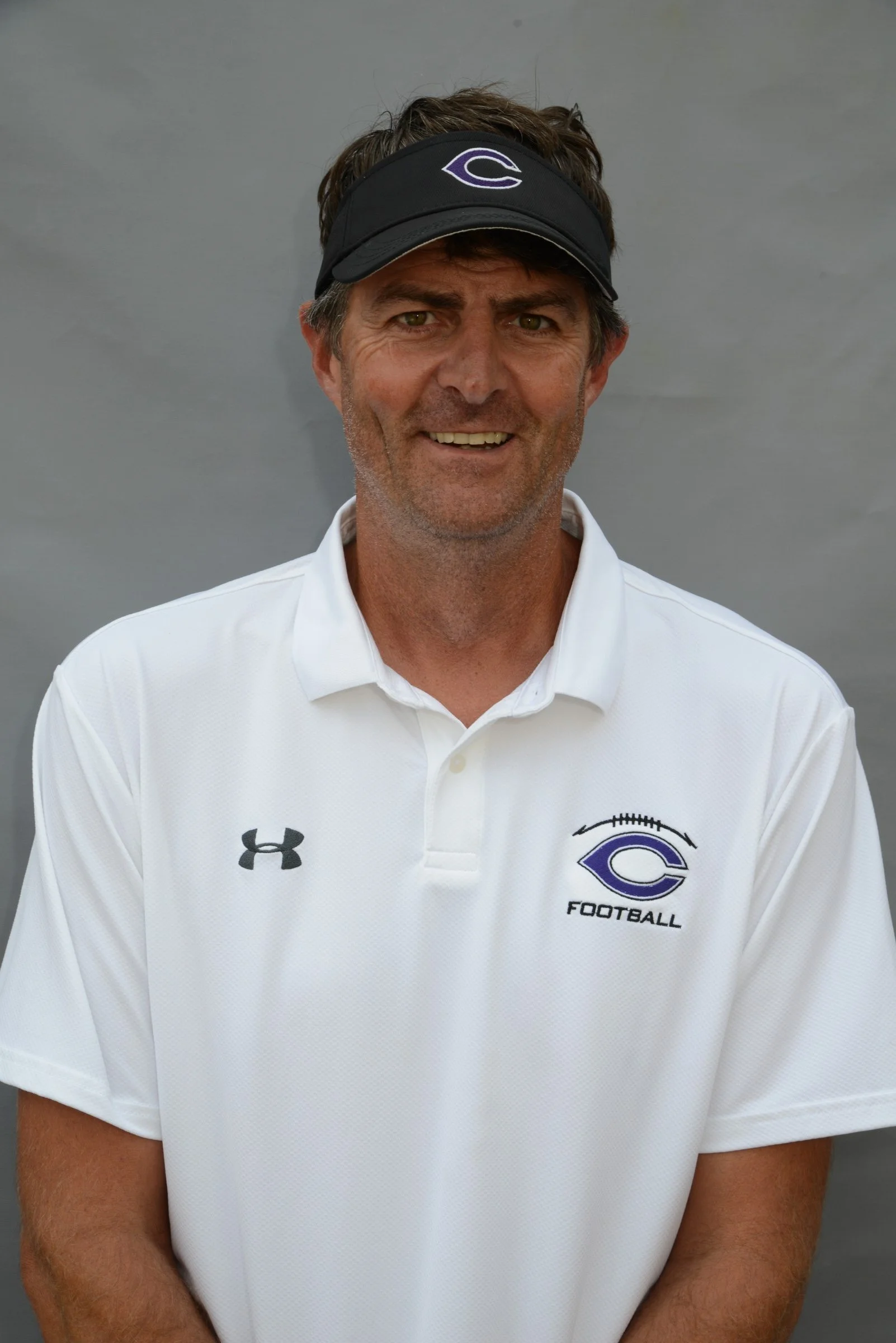 A man wearing a black visor and a white football shirt with the team logo and the word 'FOOTBALL' on it, standing against a grey wall.