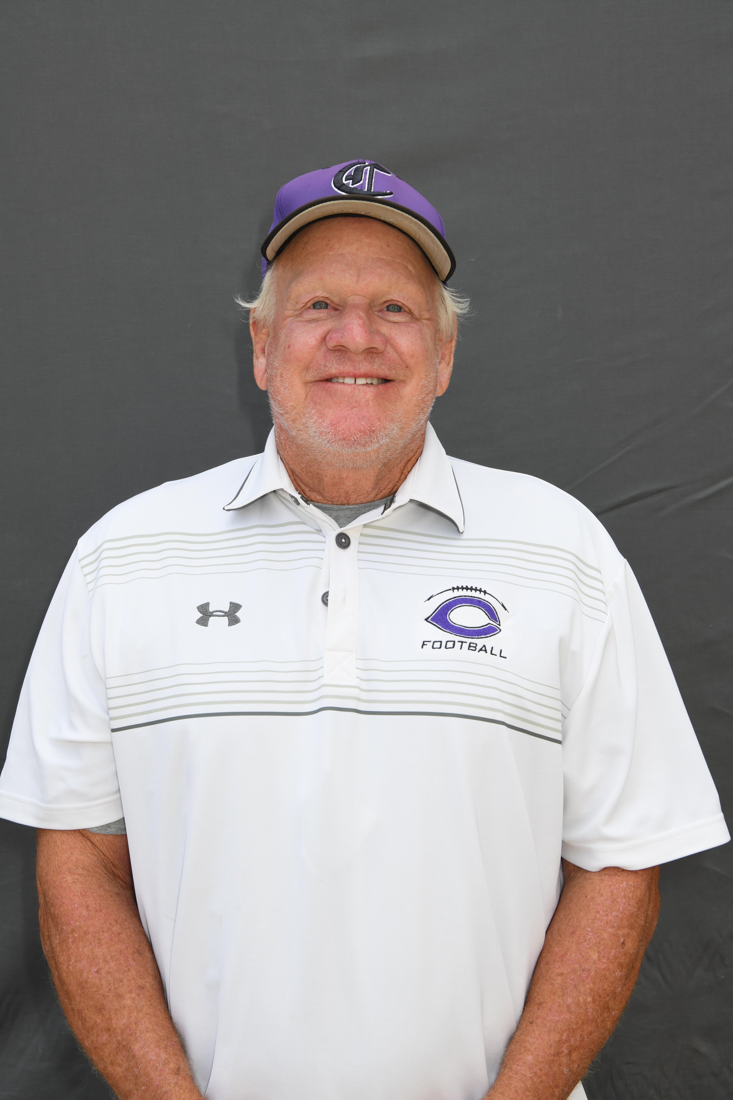 A smiling older man wearing a purple and black football cap and a white collared shirt with a football team logo and Under Armour logo, standing against a plain dark background.