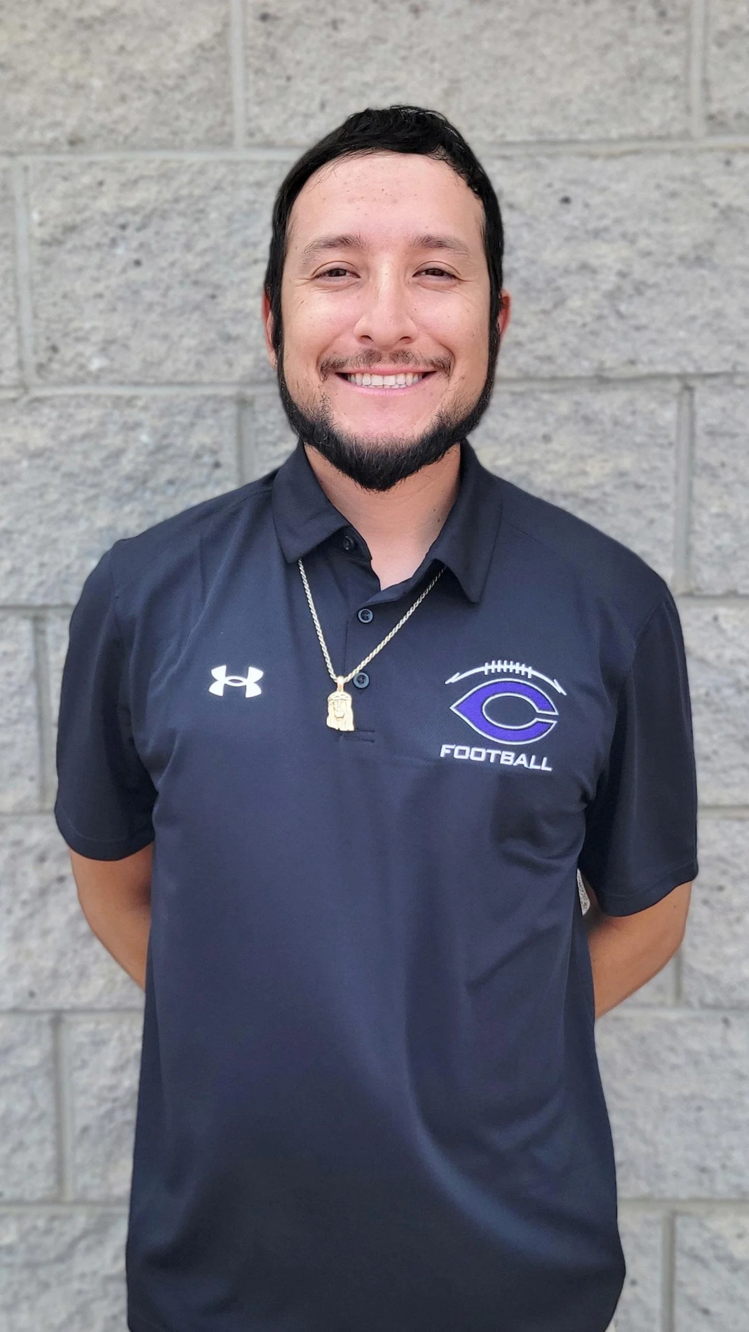 Young man with short dark hair and facial hair smiling, wearing a navy blue football team polo with logo and text, standing against a gray brick wall.