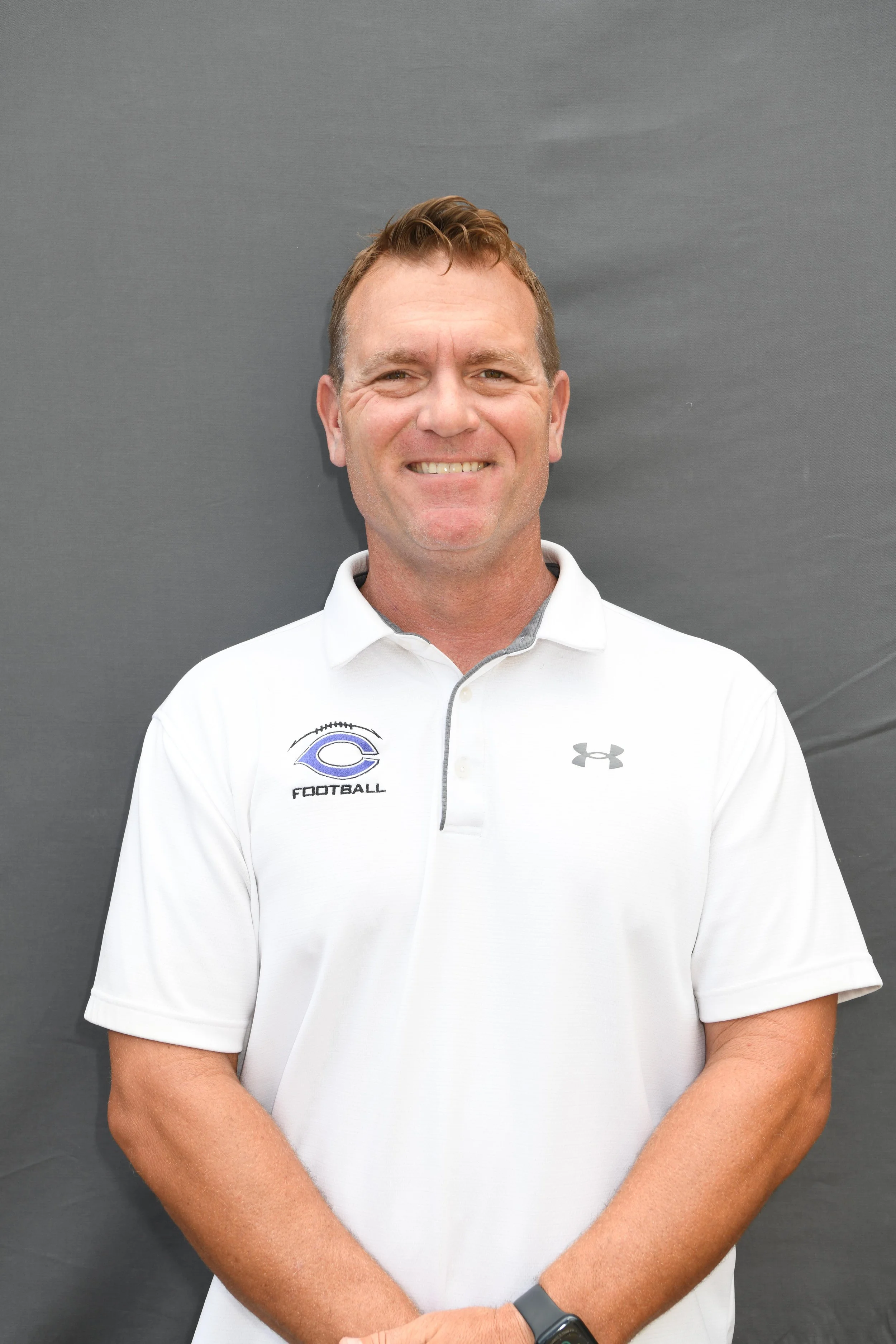 A smiling man wears a white polo shirt with a football team logo on it, standing against a plain gray background.