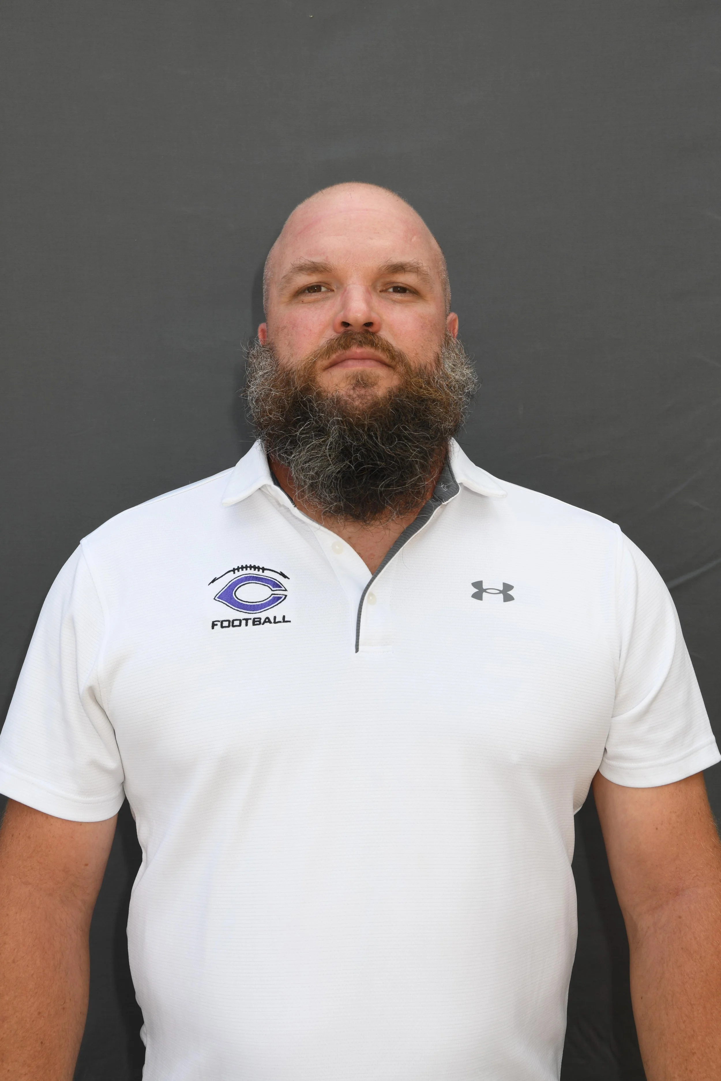 A man with a bald head and a full beard wearing a white sports polo shirt against a dark background.