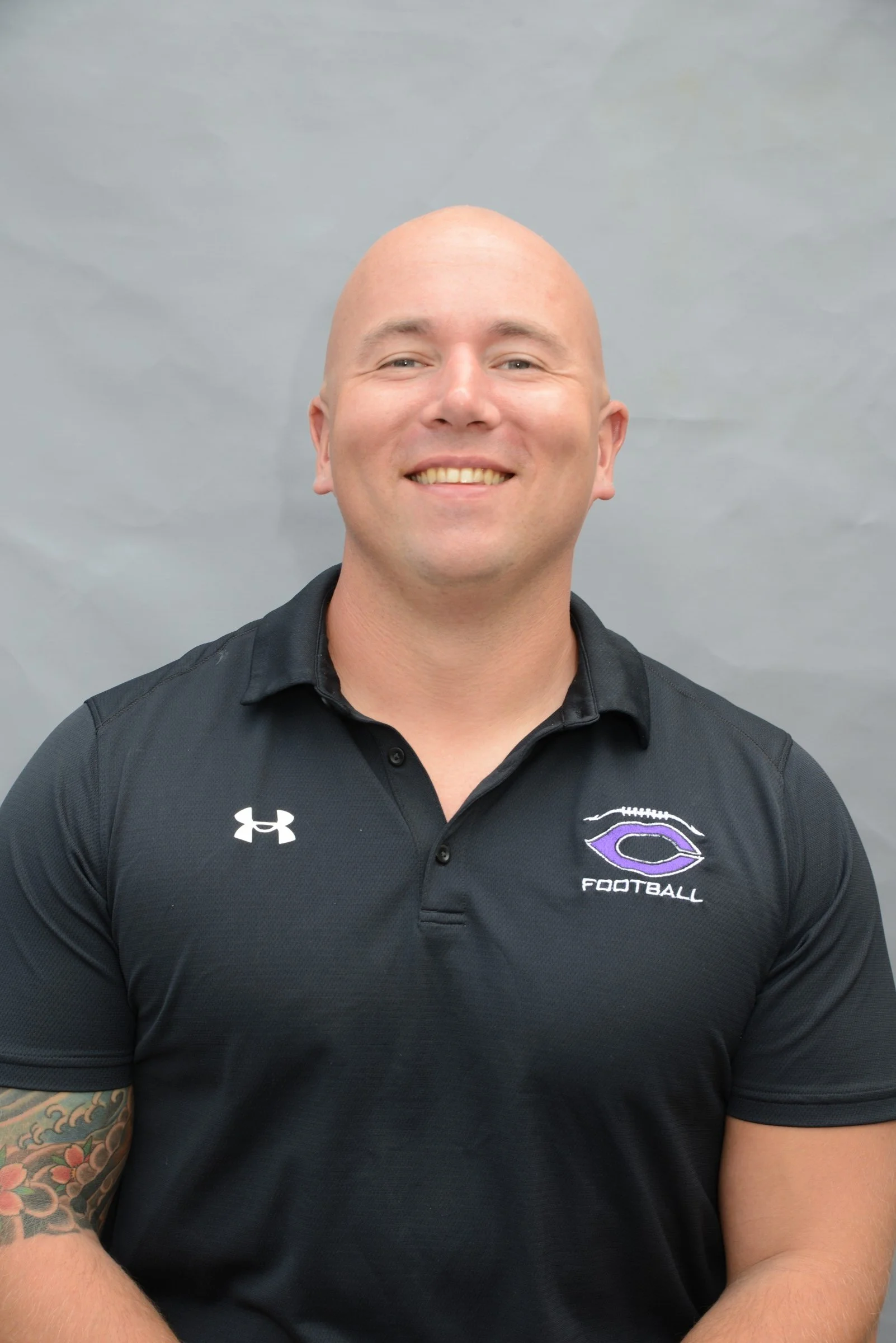 A smiling bald man wearing a black football polo shirt with the Under Armour logo and a purple football team logo on the chest, with colorful tattoos on his right arm, standing against a gray background.