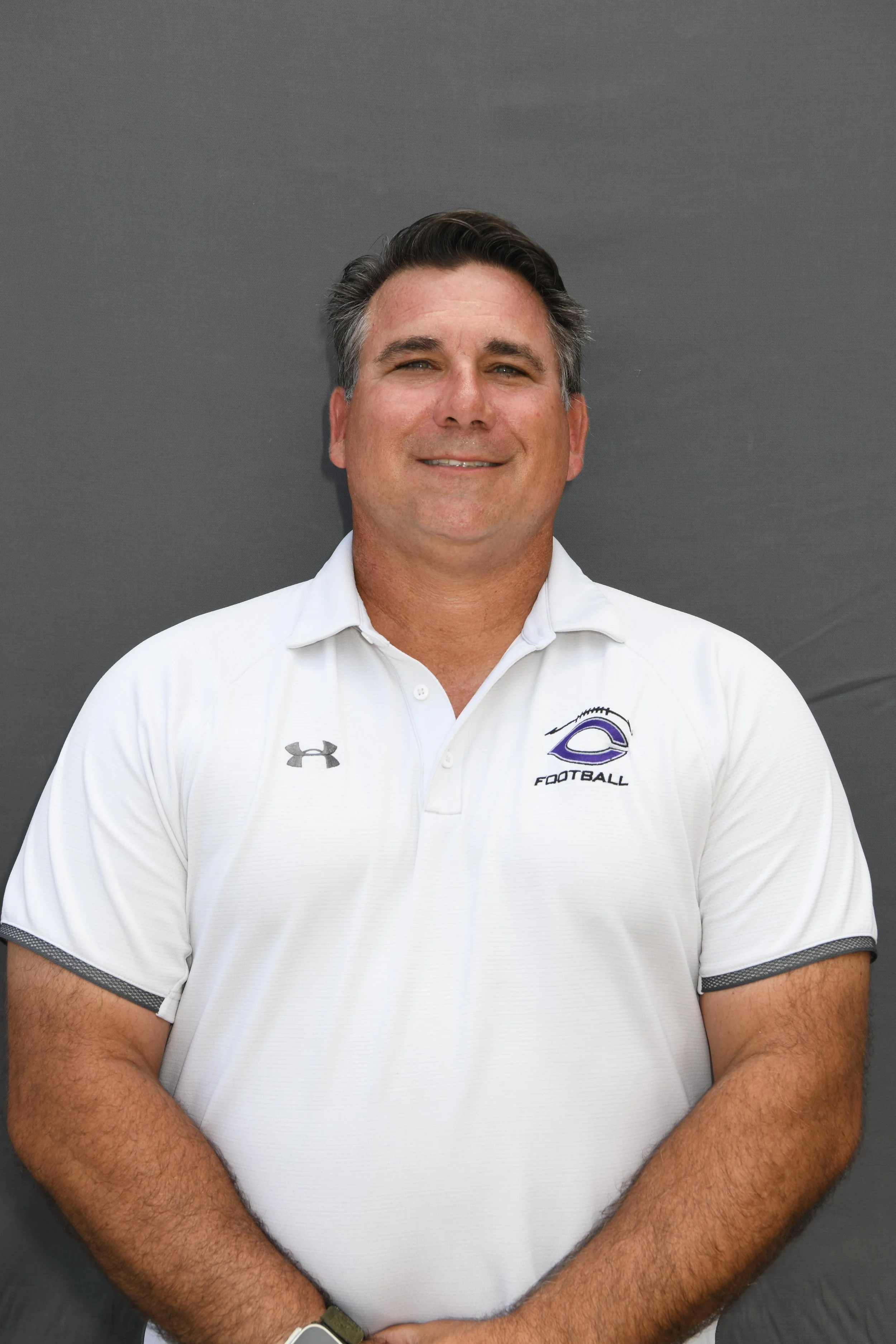 Portrait of a smiling man wearing a white polo shirt with football and Under Armour logos, standing against a gray background.