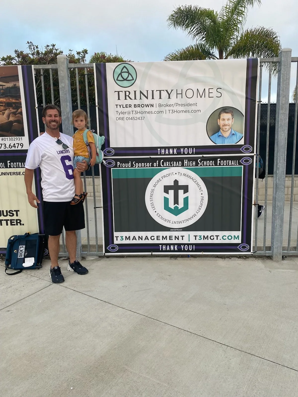 Man holding a young girl standing next to a sponsor banner at an outdoor event. The man is wearing a white sports shirt with purple lettering and black shorts, and the girl is in a yellow shirt with a backpack. The banner advertises Trinity Homes and T3 Management, with a logo, photographs, and contact information.