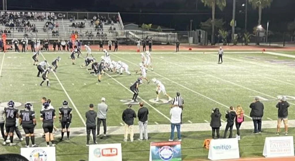 An American football game at night, with players lined up on the field, spectators in the stands, and coaches and officials on the sidelines.