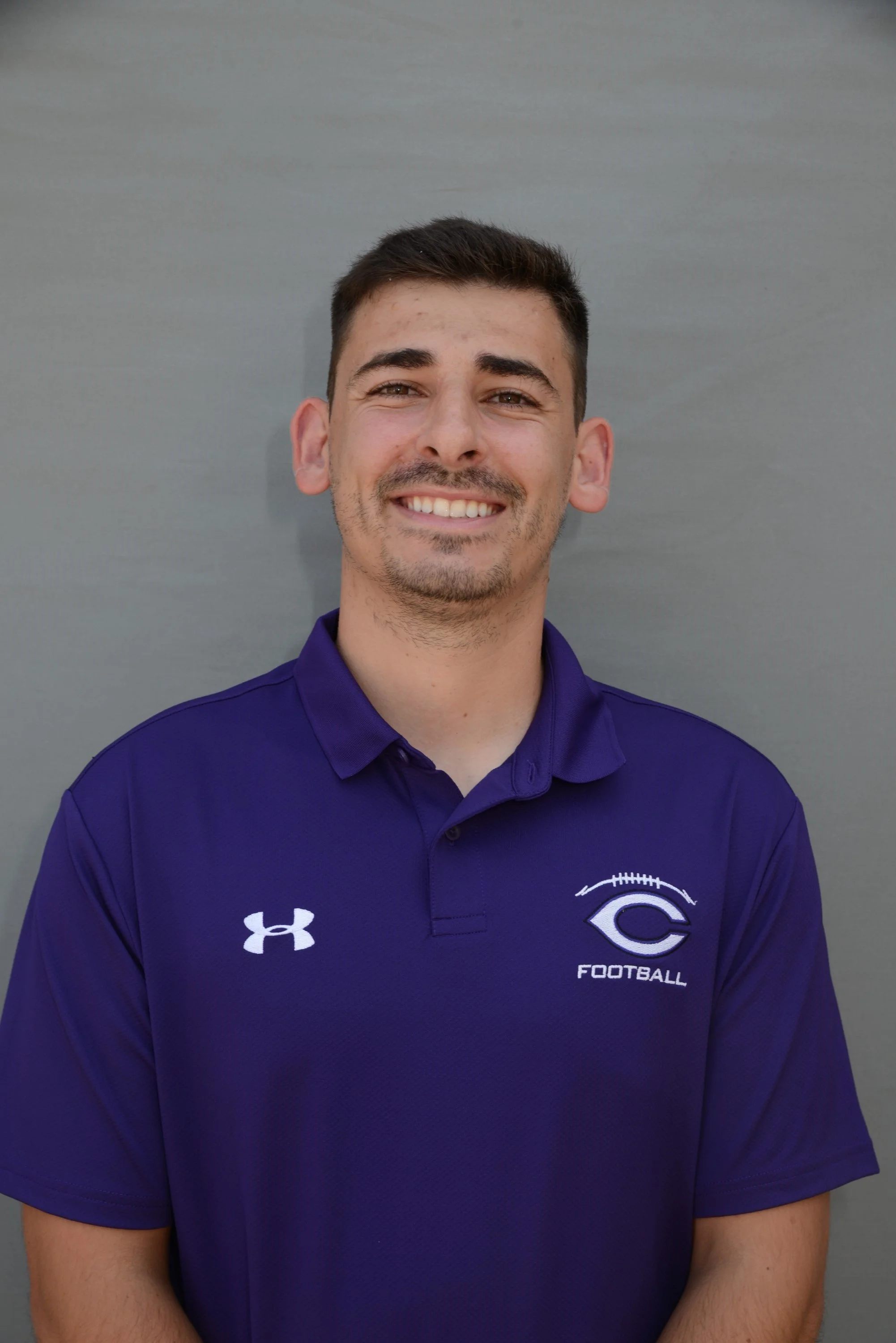 Portrait of a young man with short dark hair, light facial hair, smiling, wearing a purple football polo shirt with a logo on the left chest that says 'FOOTBALL' and the Under Armour logo on the right.