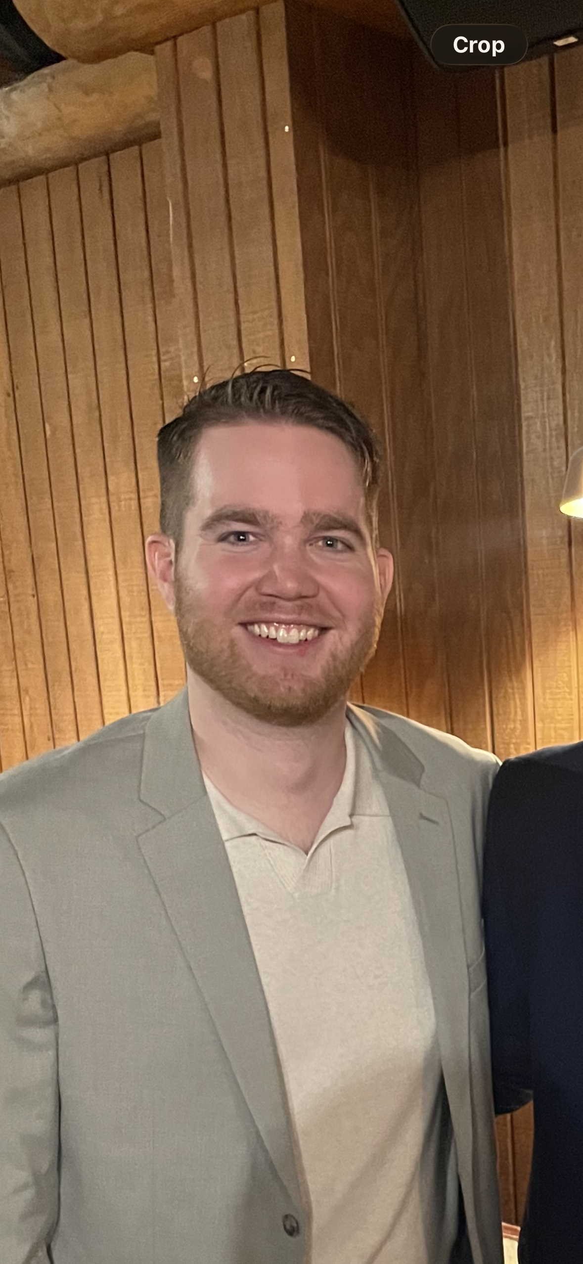 A smiling man with short brown hair, a beard, and light skin, wearing a light gray blazer over a cream-colored sweater, standing indoors in front of a wooden paneled wall.