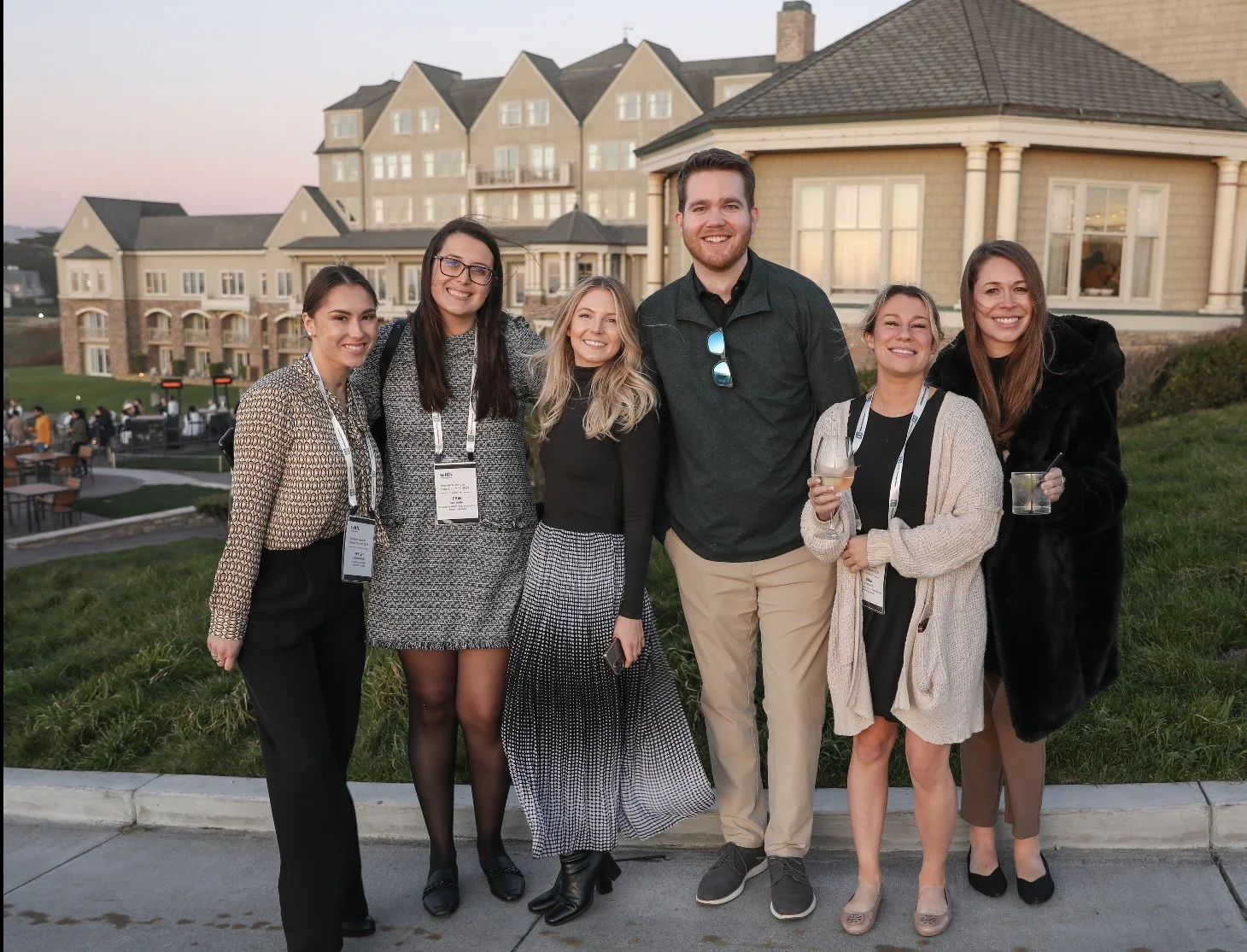 Group of six people smiling outdoors during evening at a social event, standing in front of a large residential building.