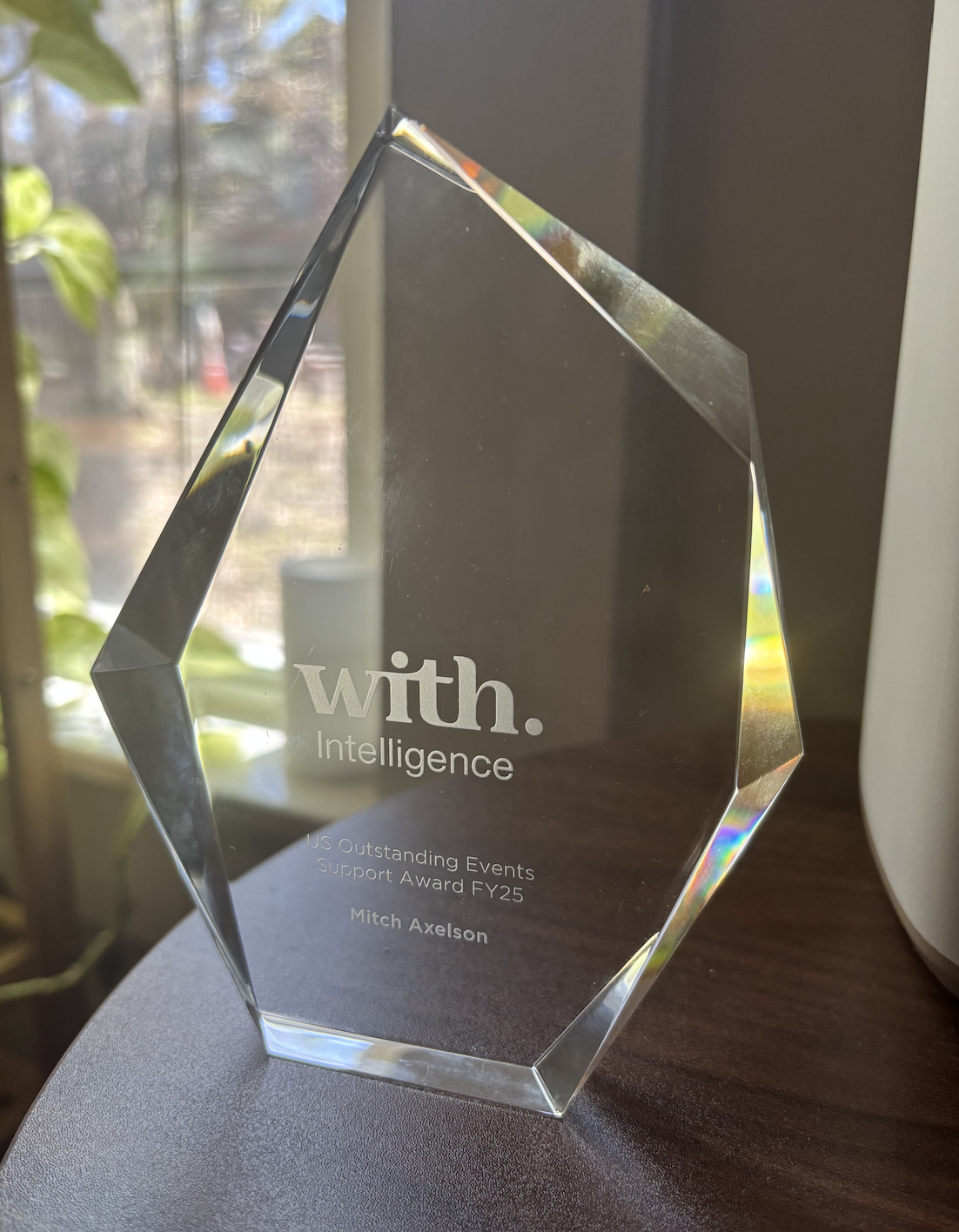 A transparent crystal award with a diamond shape, inscribed with the words 'with. Intelligence', 'US Outstanding Events Support Award FY25', and 'Mitch Axelson', placed on a dark surface near a window.