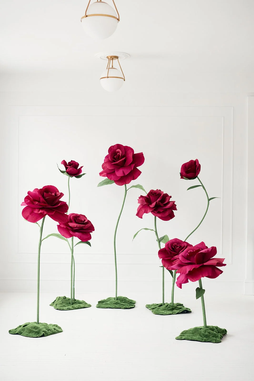 Artificial red roses with long green stems and green leaves, set on green moss bases, standing indoors with white walls and ceiling lights.