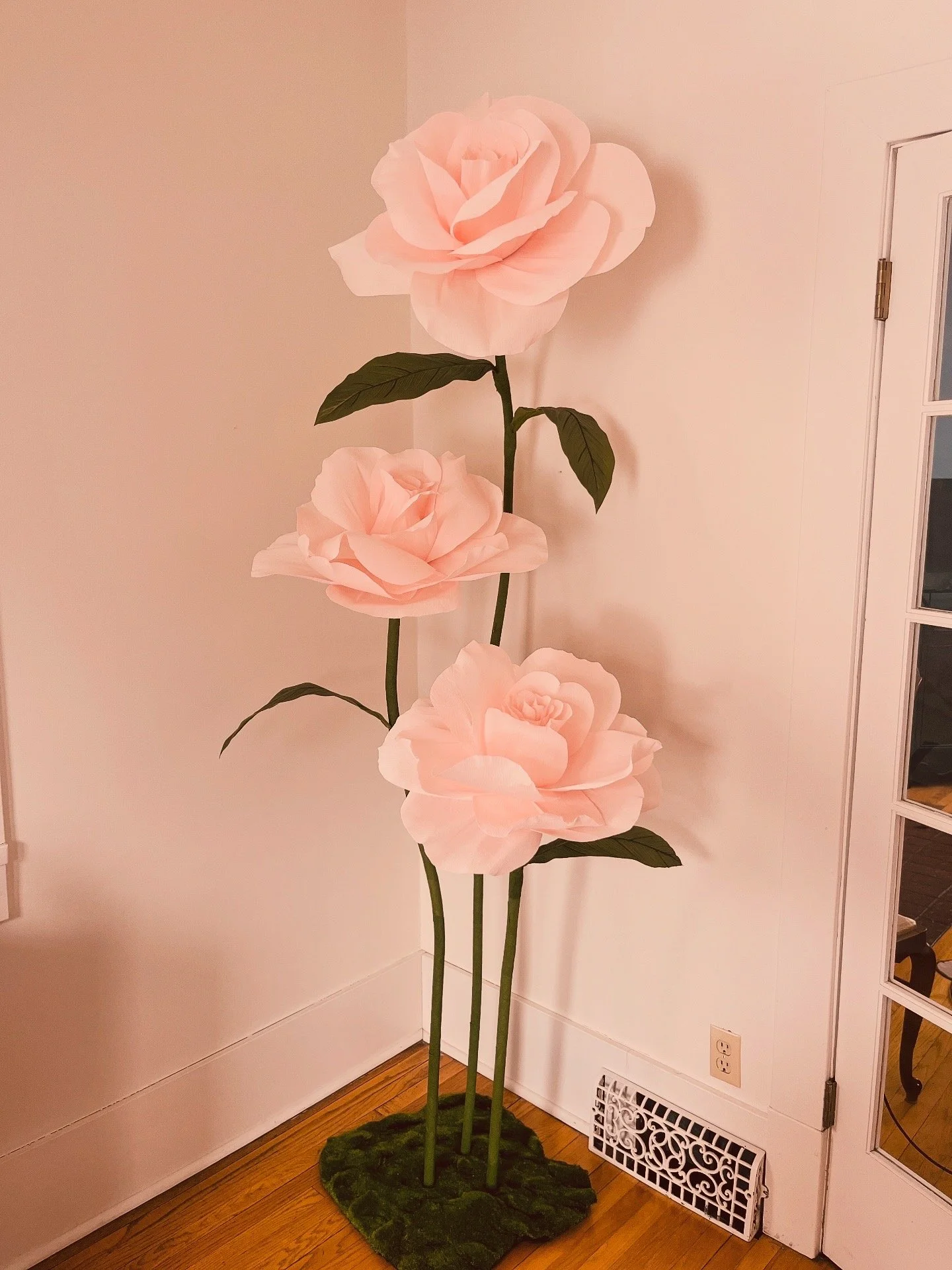 Large artificial pink roses on tall green stems with leaves, placed indoors against white walls and hardwood floors.