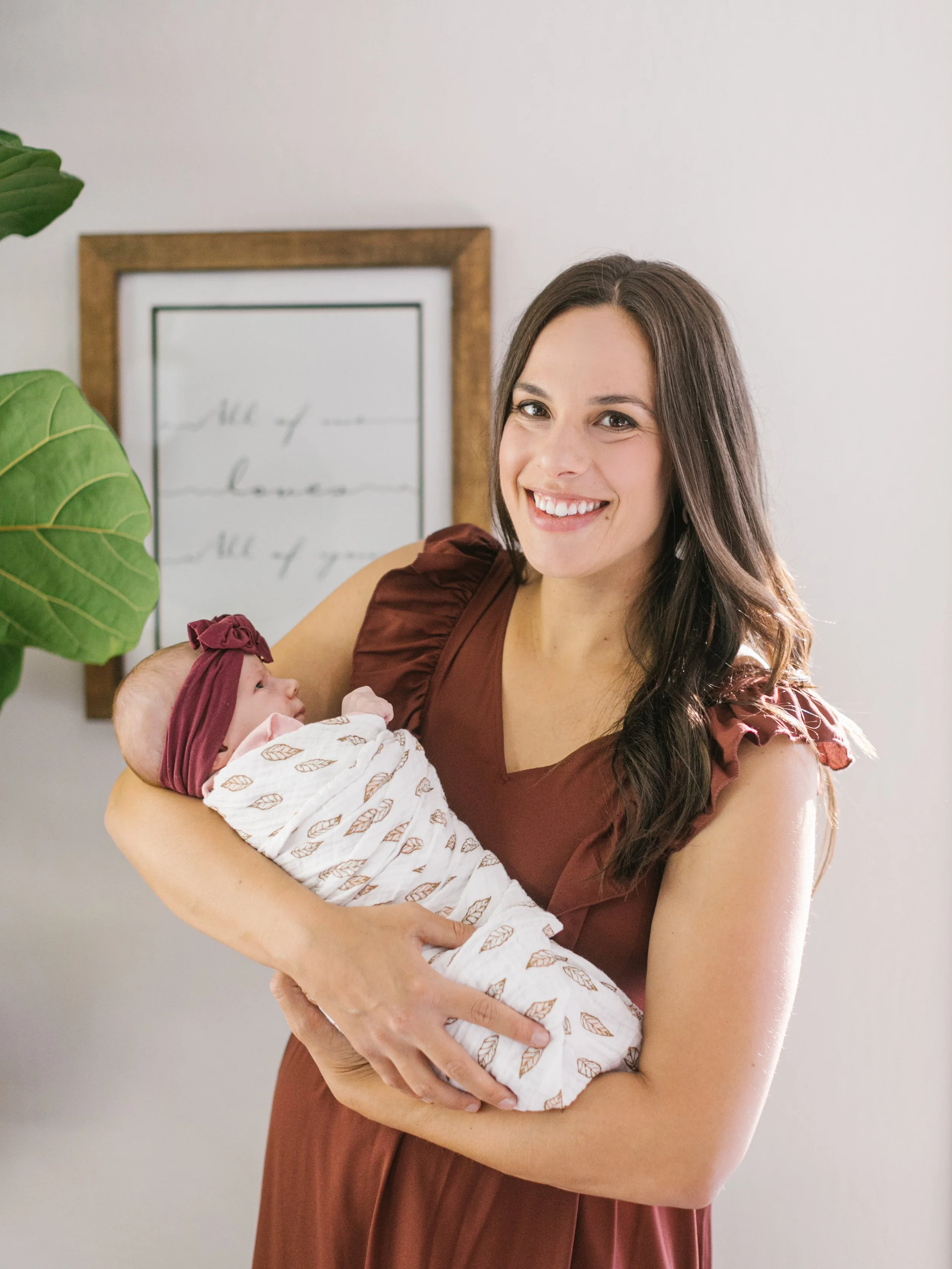 A woman with long brown hair smiling while holding a baby dressed in a patterned white outfit and a maroon headband. There is a framed artwork or sign on the wall behind them and a large green plant on the left.