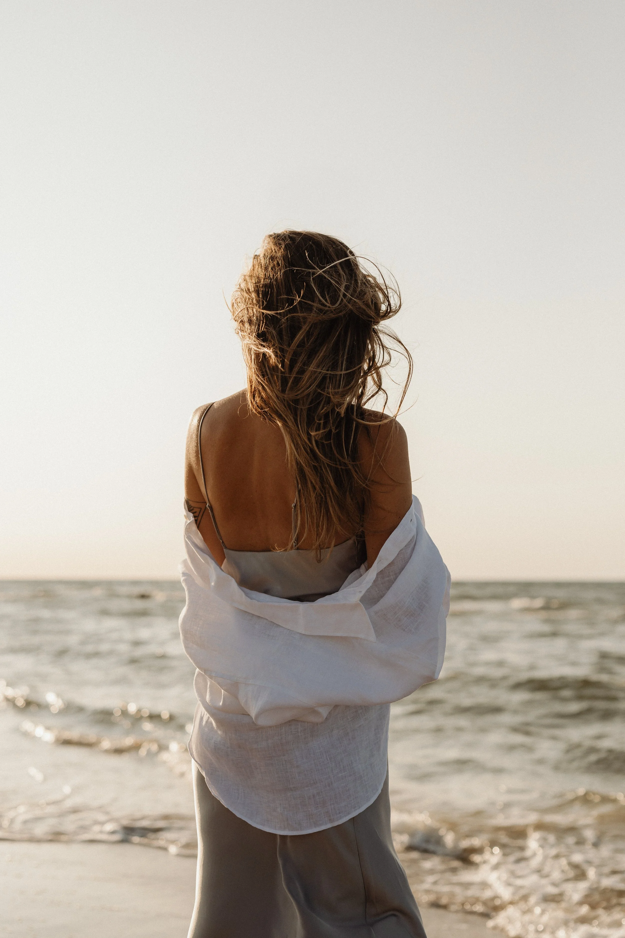 A woman with long, wavy hair standing on a beach, facing the ocean, with a light-colored button-up shirt wrapped around her waist during sunset.