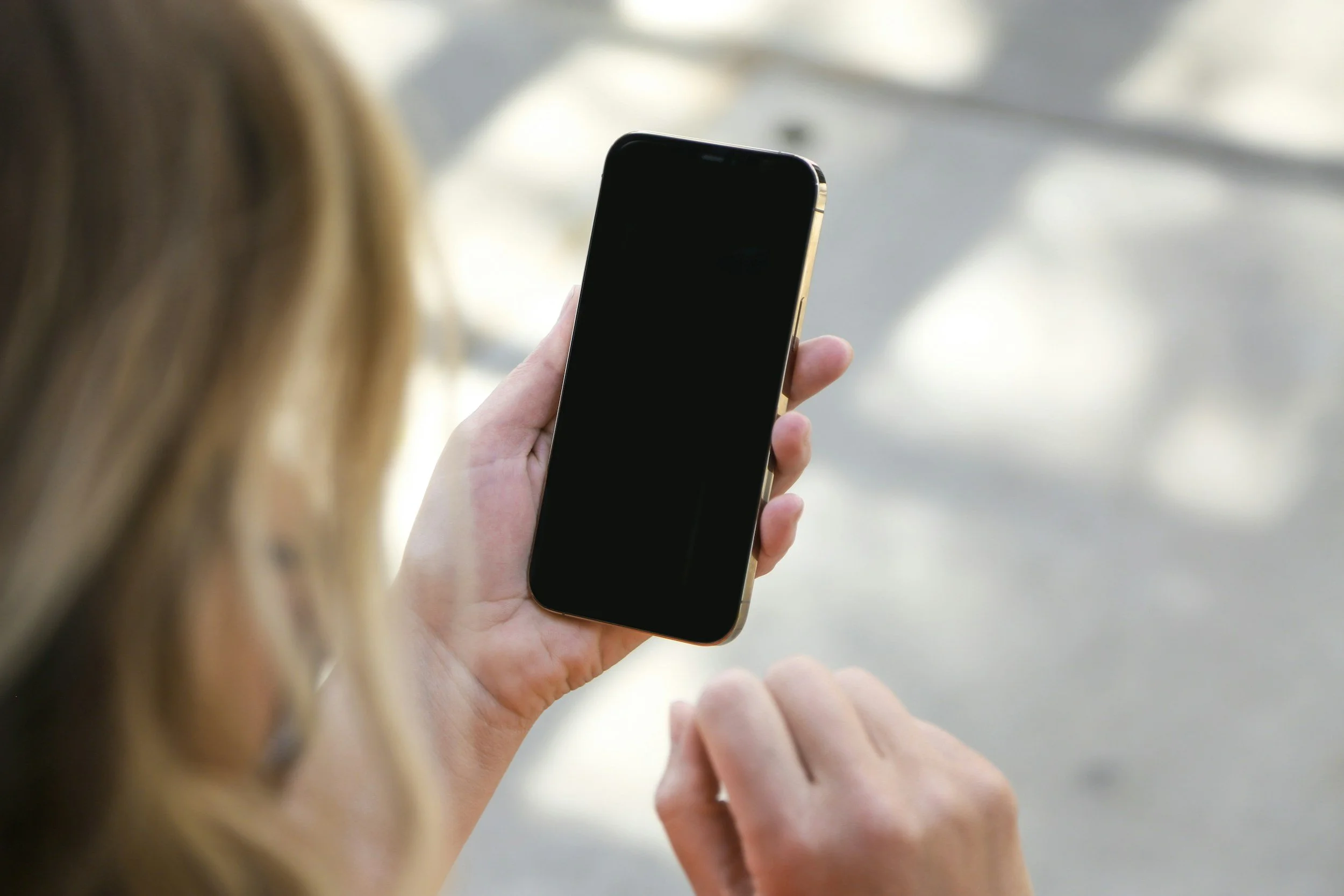 A person holding a black smartphone with a gold edge in their left hand, with the screen turned off. The person's right hand is pointing towards the phone screen. The background is blurred with a light-colored setting.