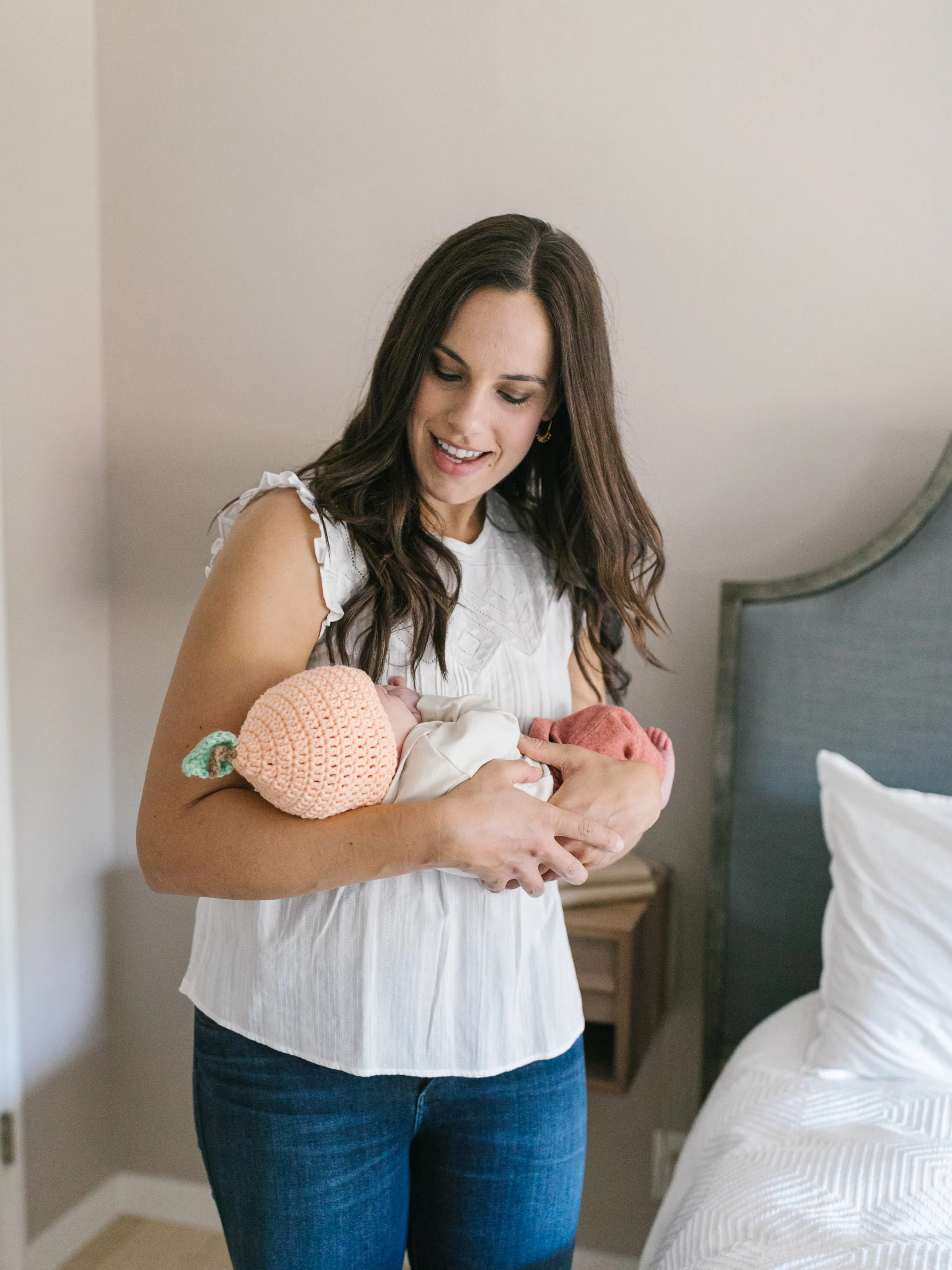 A woman with long brown hair smiling and holding a newborn baby wrapped in a blanket in a bedroom.