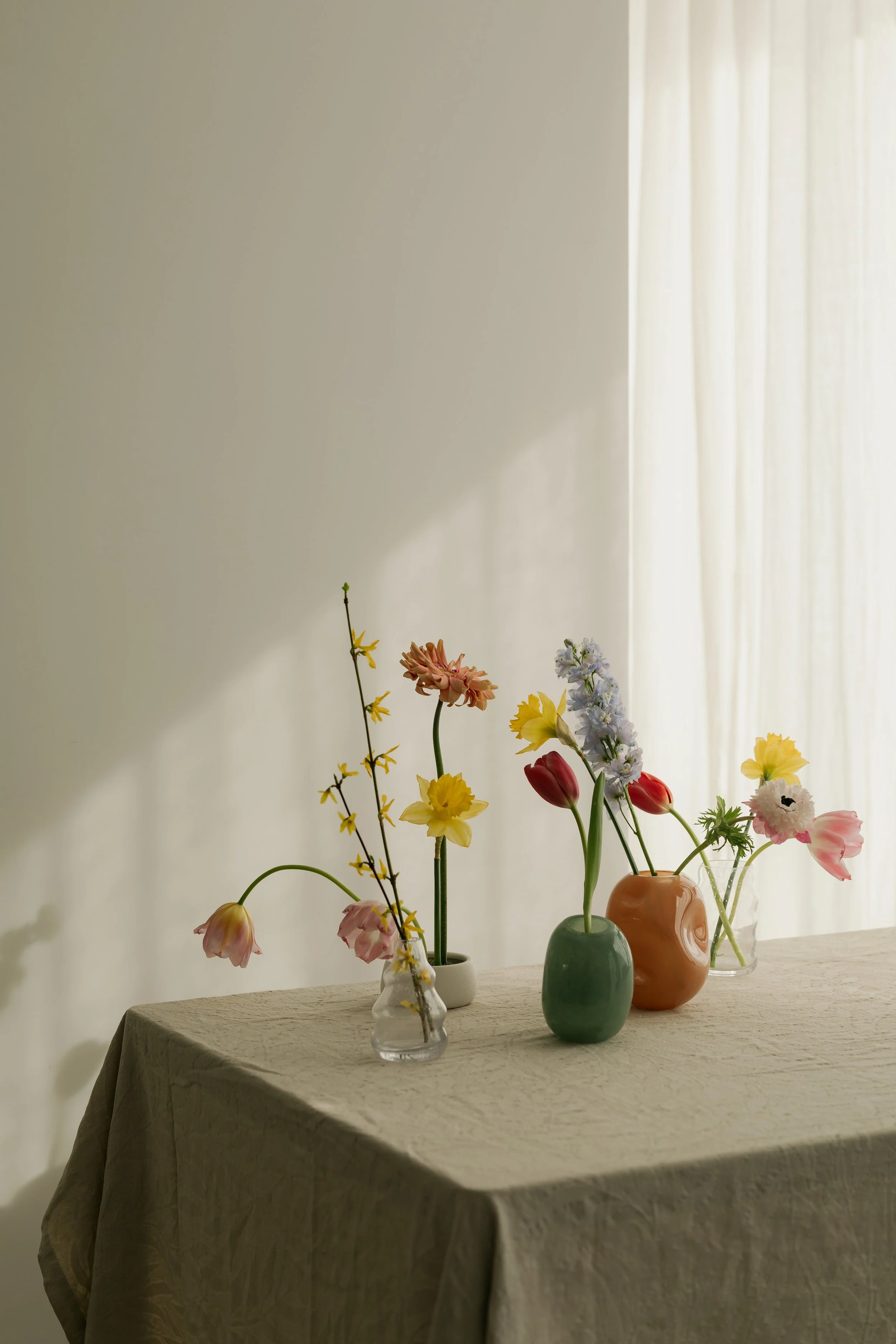 A table with a beige tablecloth holds seven vases with various flowers, set against a background of light-colored curtains.
