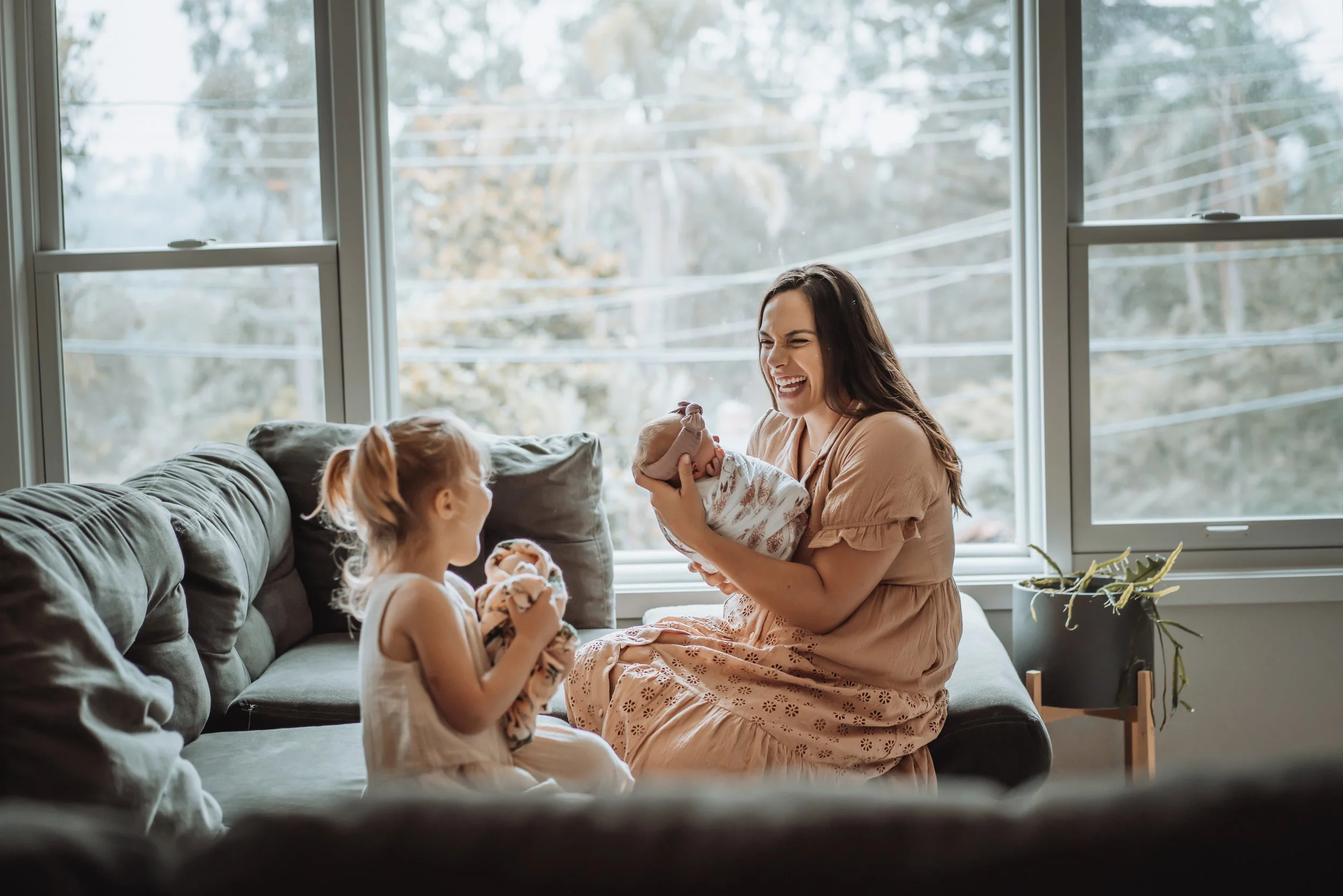 A woman with long brown hair, wearing a tan dress, sitting on a couch, holding a baby wrapped in a blanket, smiling and interacting with a young girl with red hair, also holding a doll, in a living room with large windows and a plant.