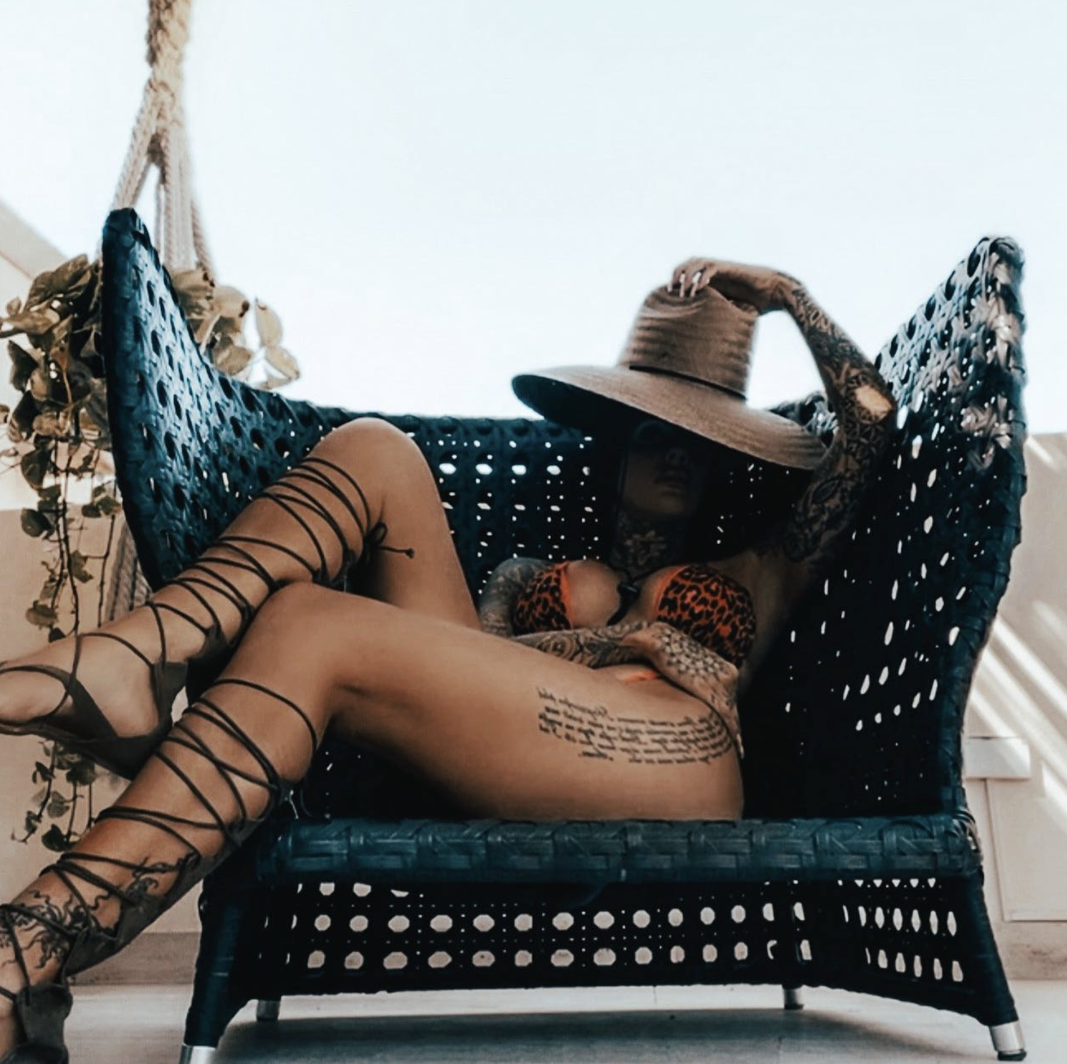 Person lounging on a black woven outdoor sofa, wearing lace-up gladiator sandals, a leopard print bra, has tattoos, and a wide-brimmed straw hat covering their face, with wine glasses nearby, and dried plants hanging on the wall.