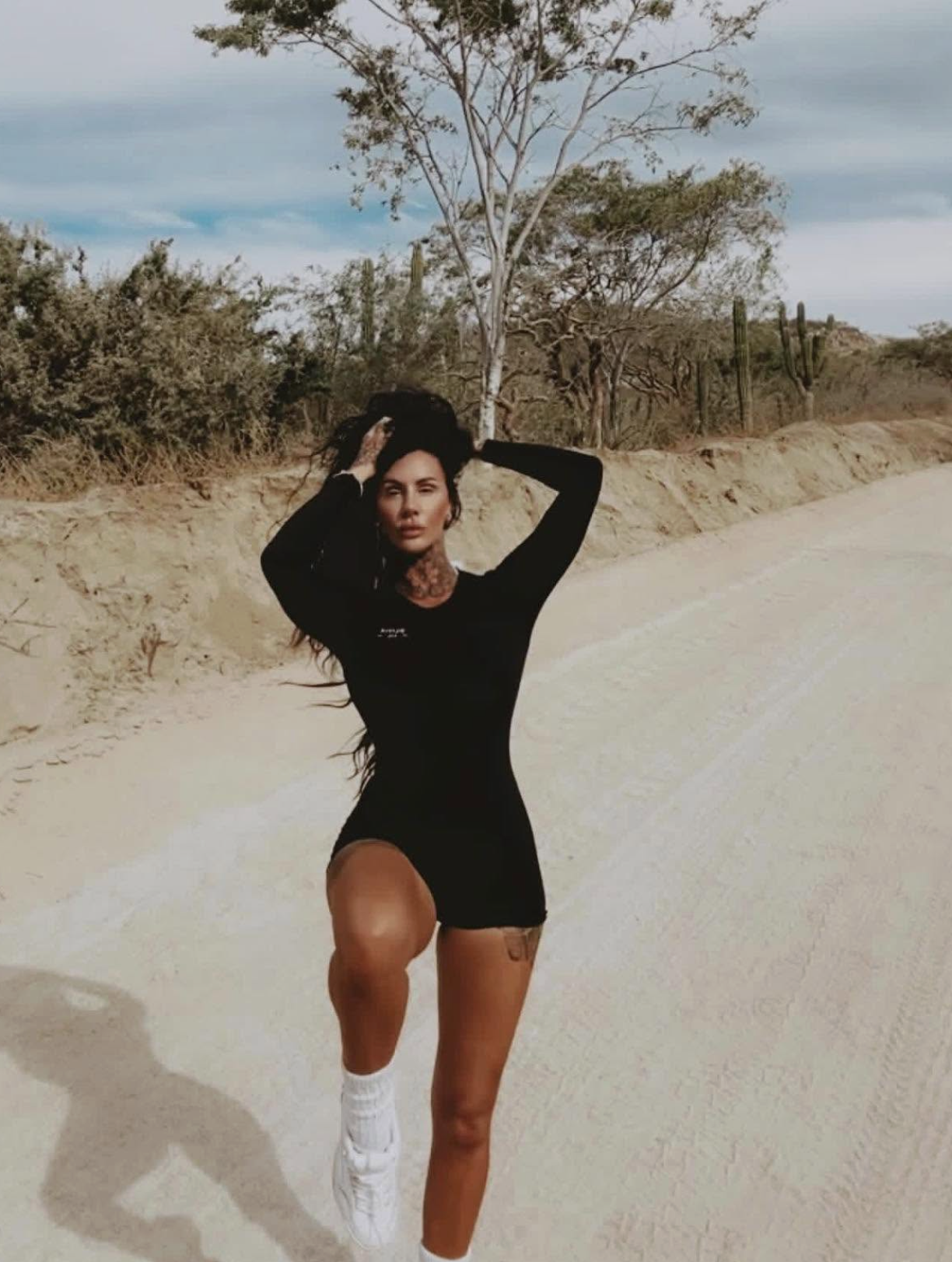 A woman with tattoos on her neck and legs is standing on a dirt road in a desert landscape, wearing a black dress and white sneakers, with her arms raised above her head and one leg bent.