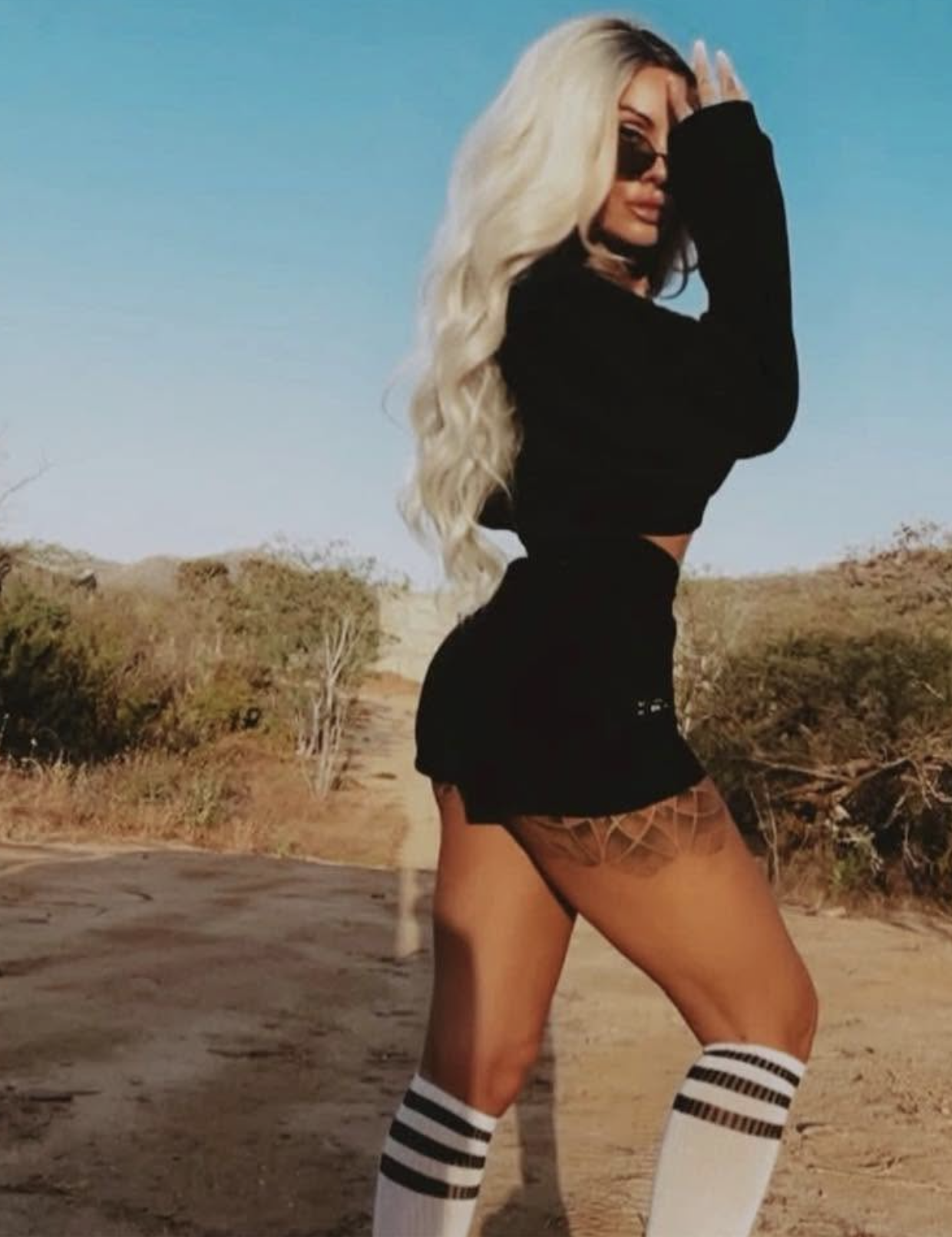 A woman with long blonde hair, wearing sunglasses, a black crop top, black shorts, and knee-high white socks with black stripes, posing outdoors in a desert landscape.
