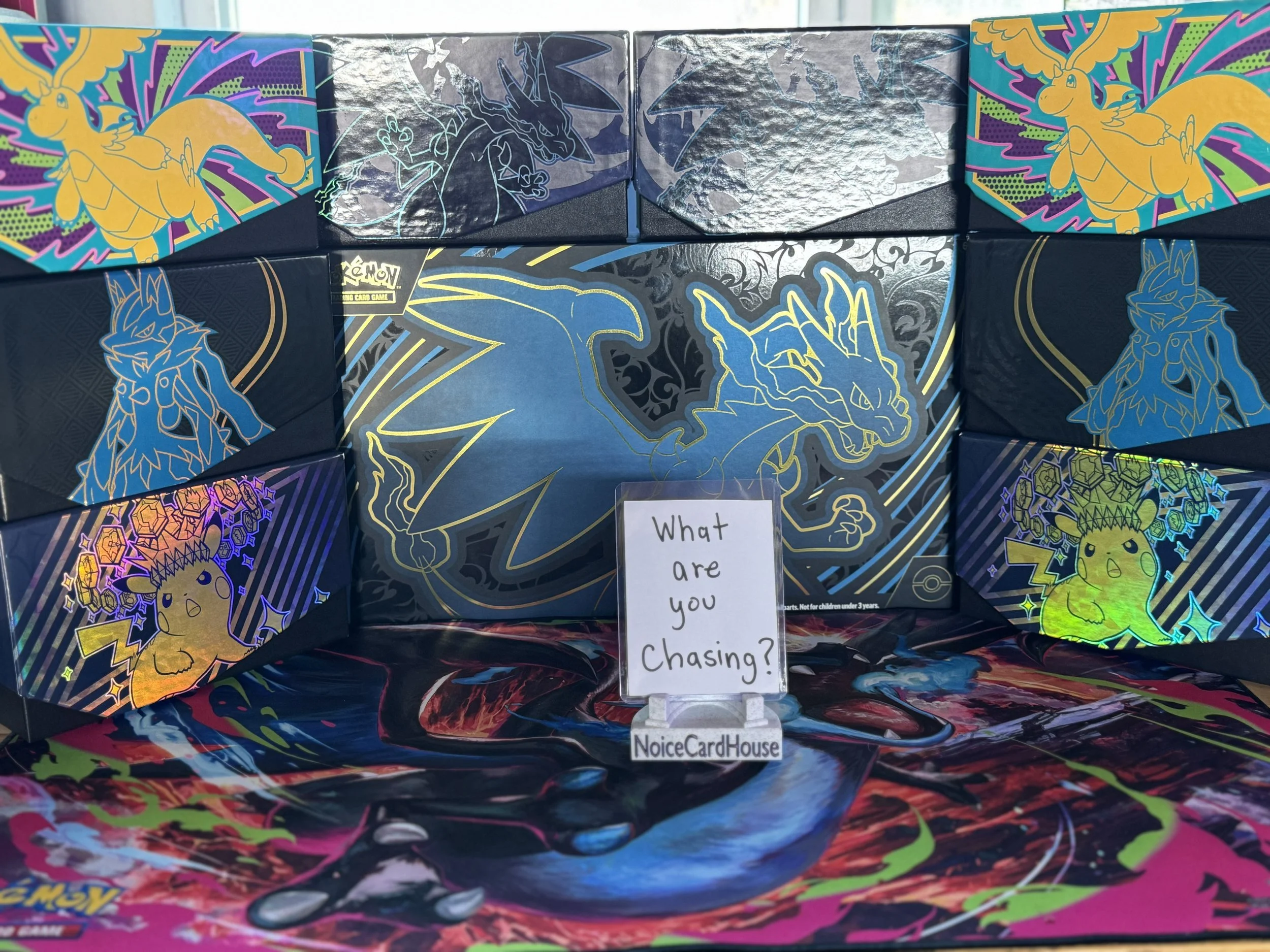 Colorful collage of Pokémon-themed boxes featuring Garchomp, Charizard, and Pikachu, with a handwritten sign reading 'What are you choosing? NoiceCardHouse'.