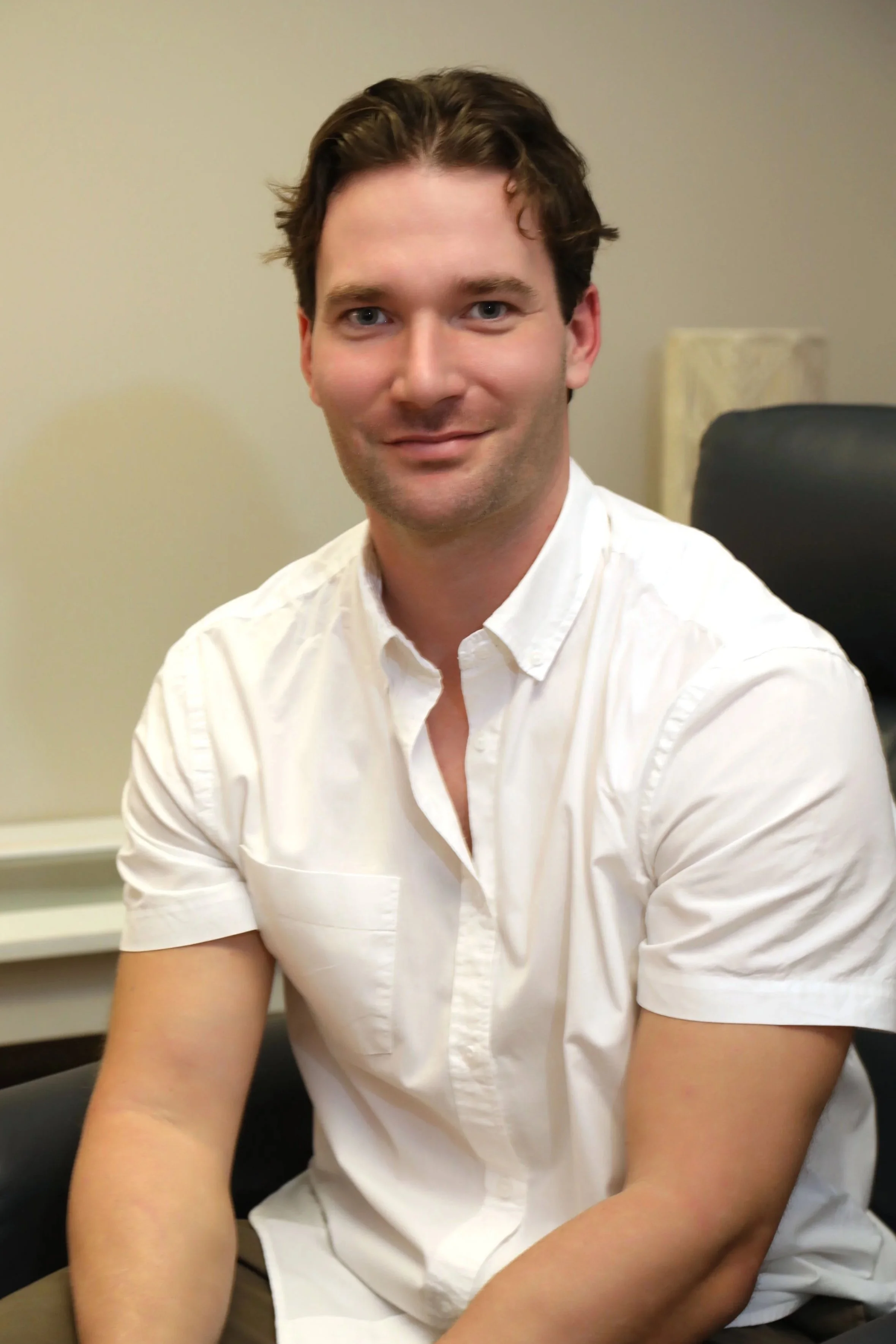 Head and shoulders portrait of a young man with light skin, brown hair, wearing a white button-up shirt, sitting in an office chair in an indoor setting.