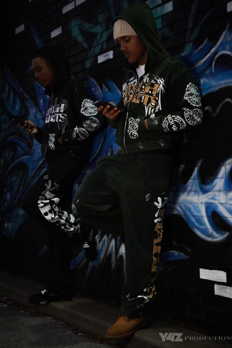 Two young men standing against a colorful graffiti wall, looking at their phones, wearing hooded sweatshirts and matching pants with graphic prints.