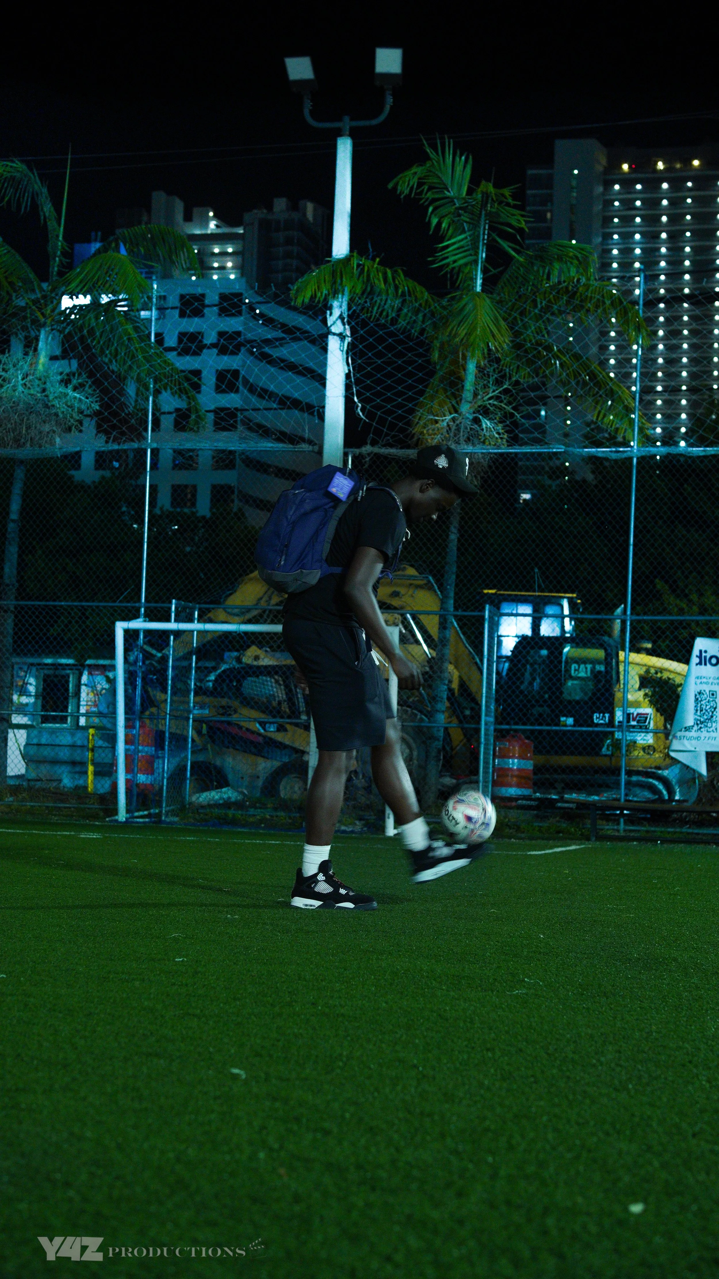A person wearing black clothing and a backpack is standing on a green artificial turf soccer field, juggling a soccer ball at night.