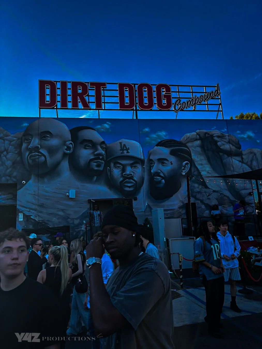 Crowd of people gathered outside Dirt Dog Compuound with a large mural of four men on the building wall, featuring a mountain landscape and the sign "Dirt Dog Compound" on top.