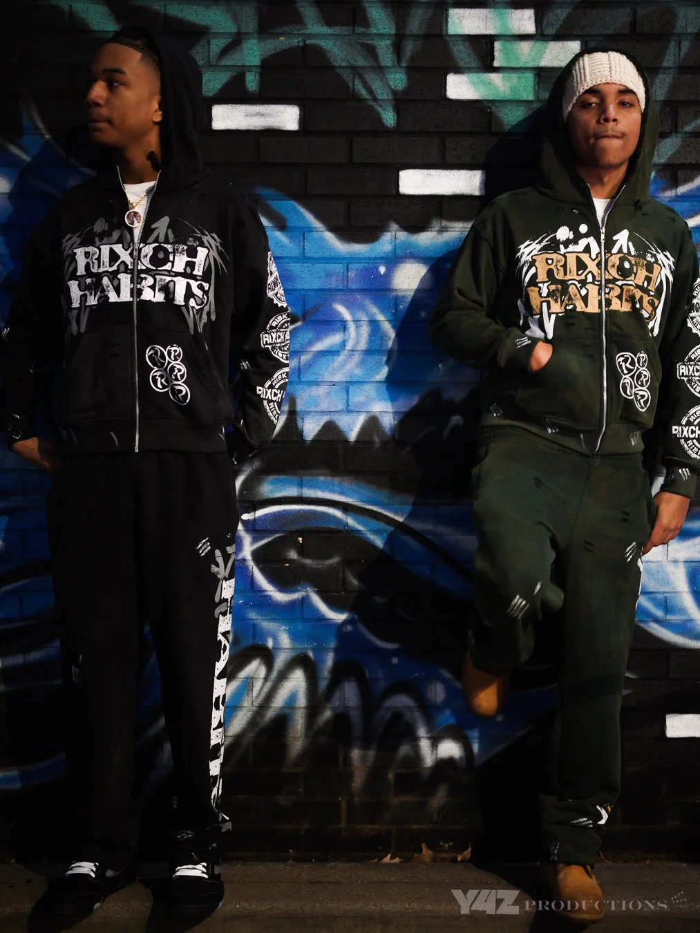 Two young brand owners in black and green hoodies with graffiti-style graphics and text, standing in front of a colorful graffiti mural on a brick wall, one with hands in pockets and wearing a beanie, the other looking away to the side.