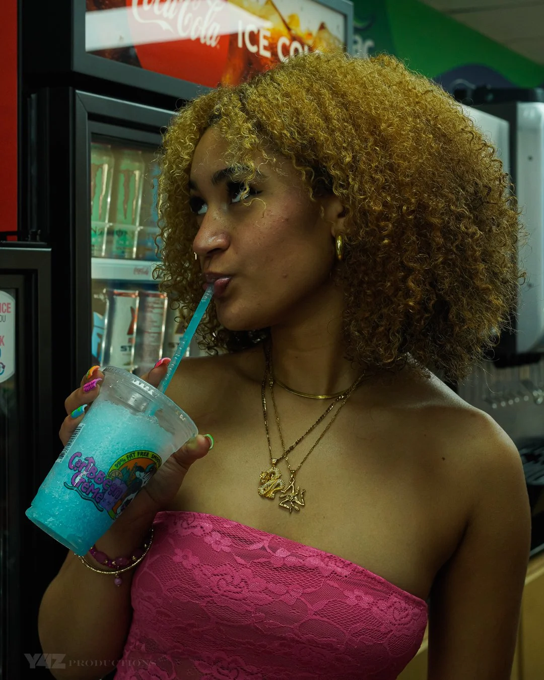 A woman with curly blonde hair drinking a blue beverage through a straw from a clear plastic cup, wearing a pink strapless lace top, gold earrings, and layered gold necklaces. She is standing in front of a refrigerator at a convenience store.