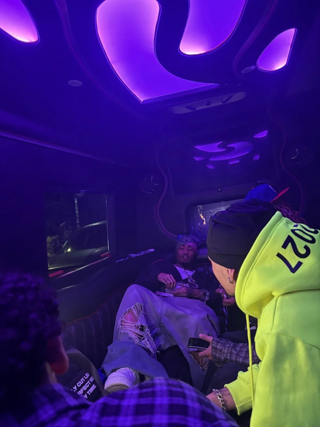 People sitting inside a bus or limo with purple and green neon lighting, one person wearing a black hoodie and a neon yellow jacket, others seated around, some taking pictures or looking at their phones.