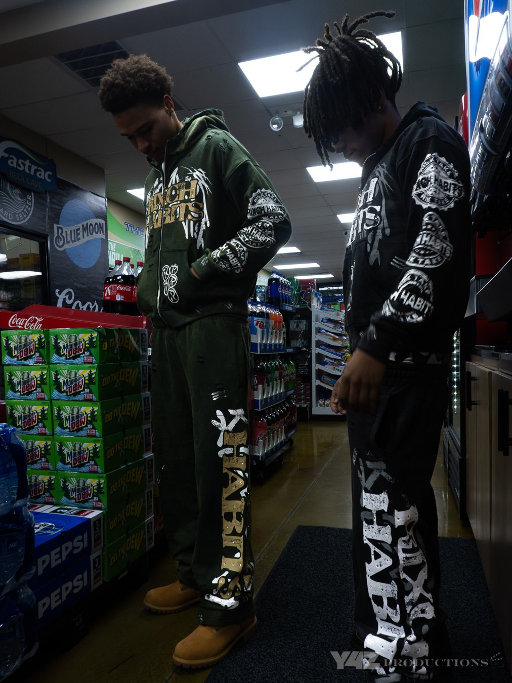Two young men standing in a convenience store aisle, wearing matching black and green hoodies and sweatpants with bold graphic text, looking down at products on the shelves.