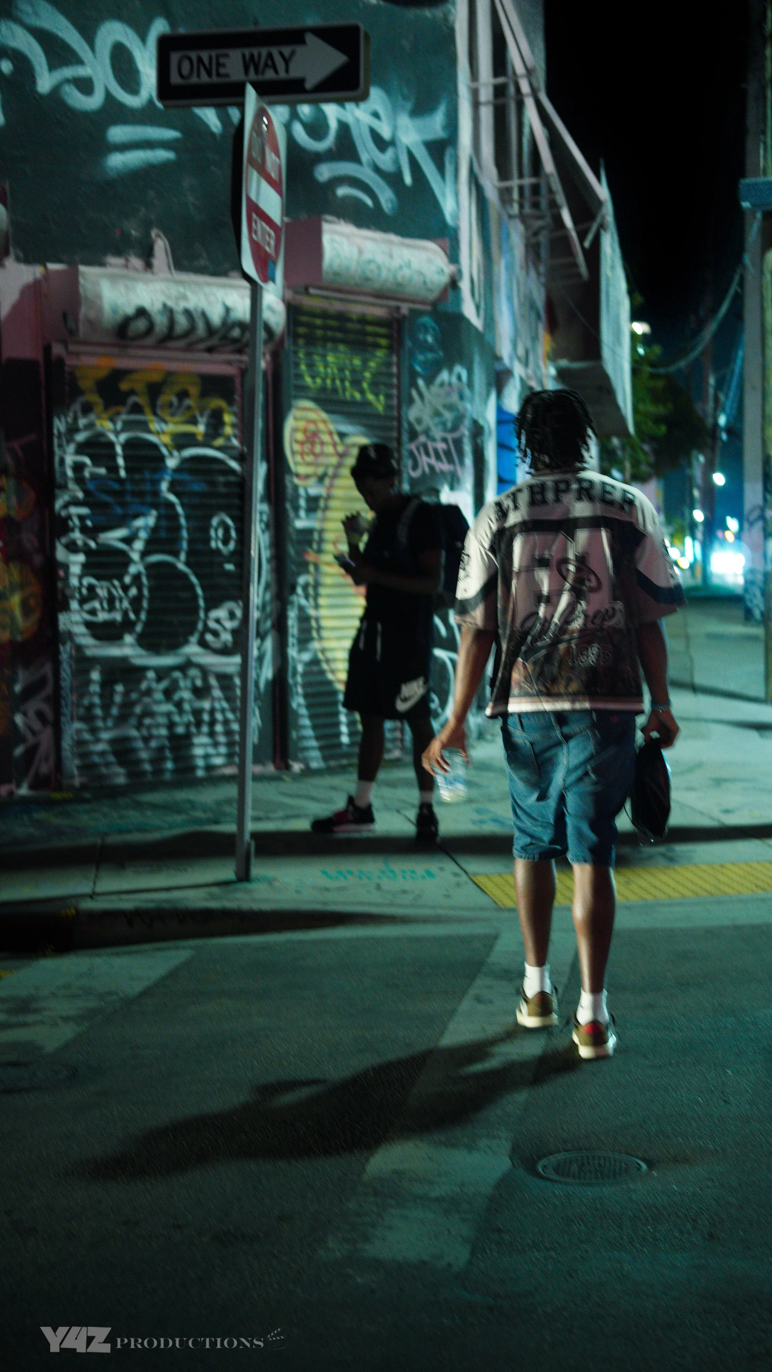 Two young men walking on a city sidewalk at night, in Miami, one is holding a water bottle, graffiti-covered building in the background, street signs, and streetlights illuminating the scene.