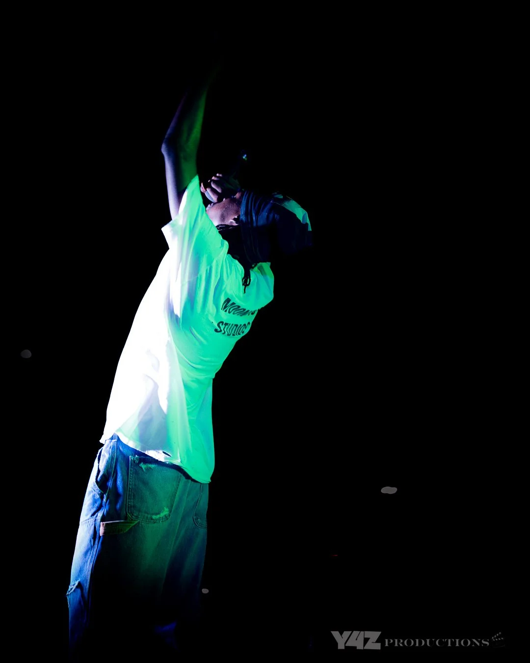 Ezcodylee  performing in a dark environment wearing a bright green and black jacket with the words "STUDIO" on it and blue jeans, holding a microphone near their mouth, with neon lighting effects.