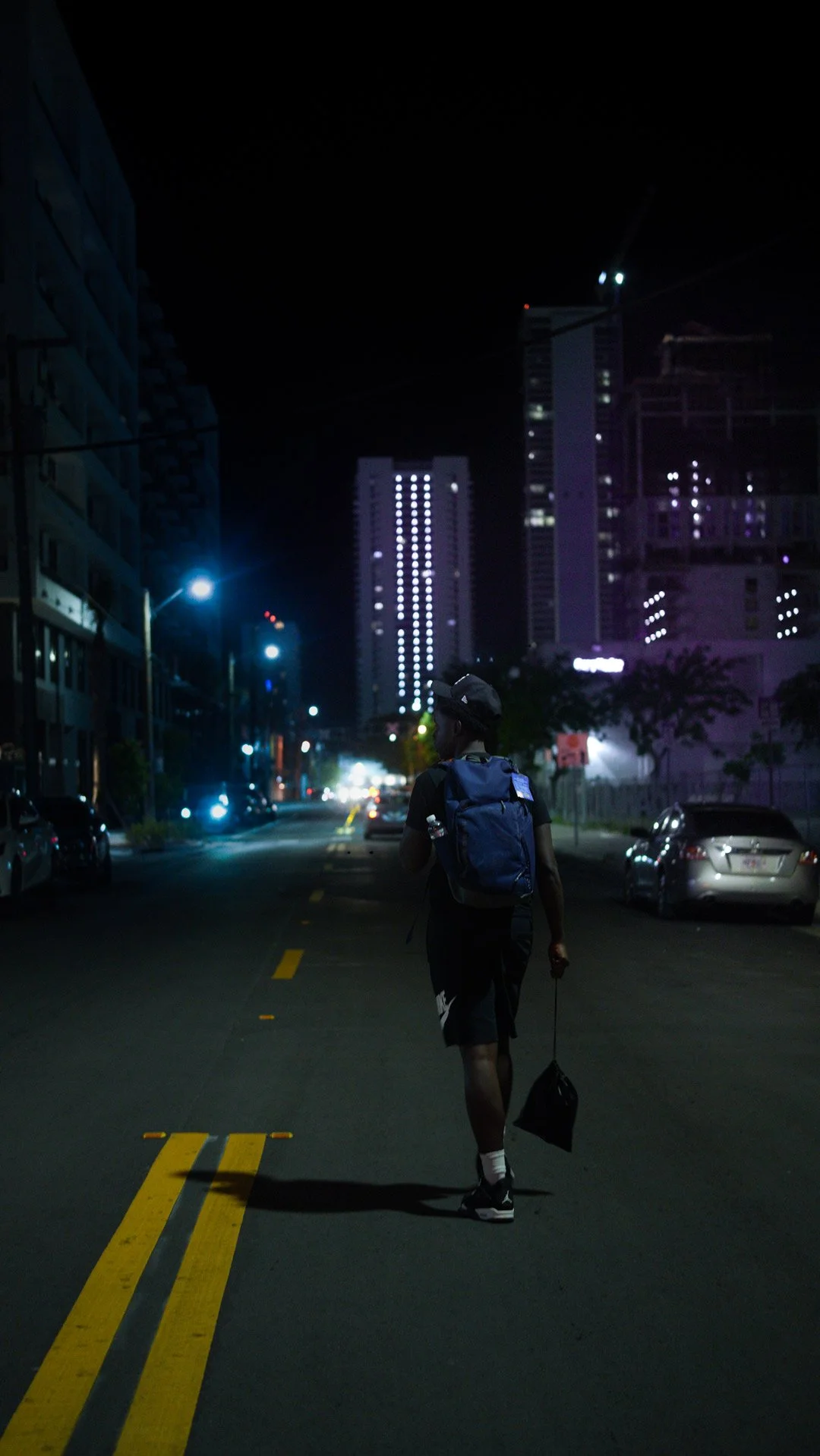A person walking alone on an empty city street at night, carrying a backpack and a small bag, with tall buildings and city lights in the background.