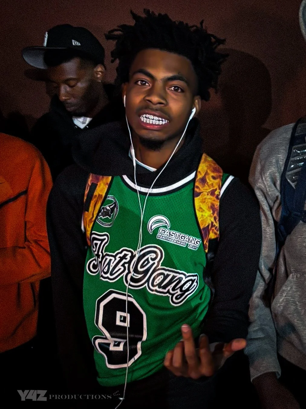lazerdim700  wearing a green and black basketball jersey, earbuds, and grinning showing teeth, standing in a group.