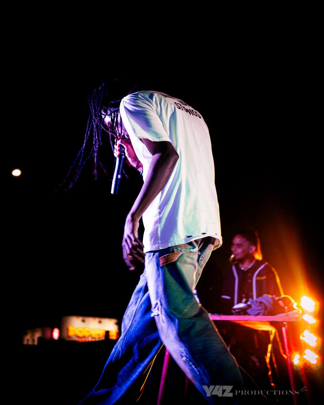 Ezcodylee  holding a microphone on stage during a night event, with colorful stage lighting and a DJ in the background.