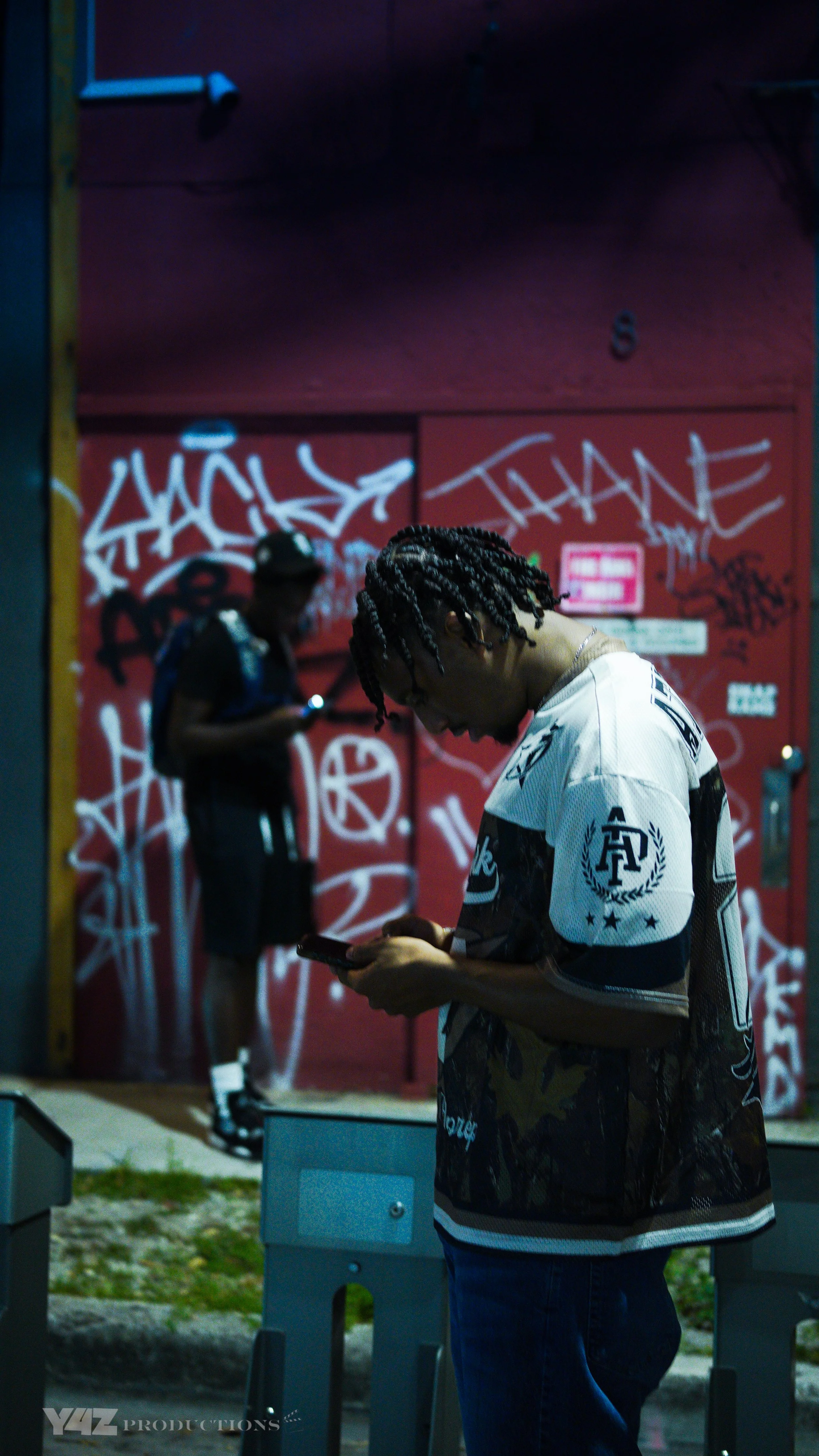 Two young men stand outside in front of a graffitied red wall, with one looking at his phone and the other looking at his phone.