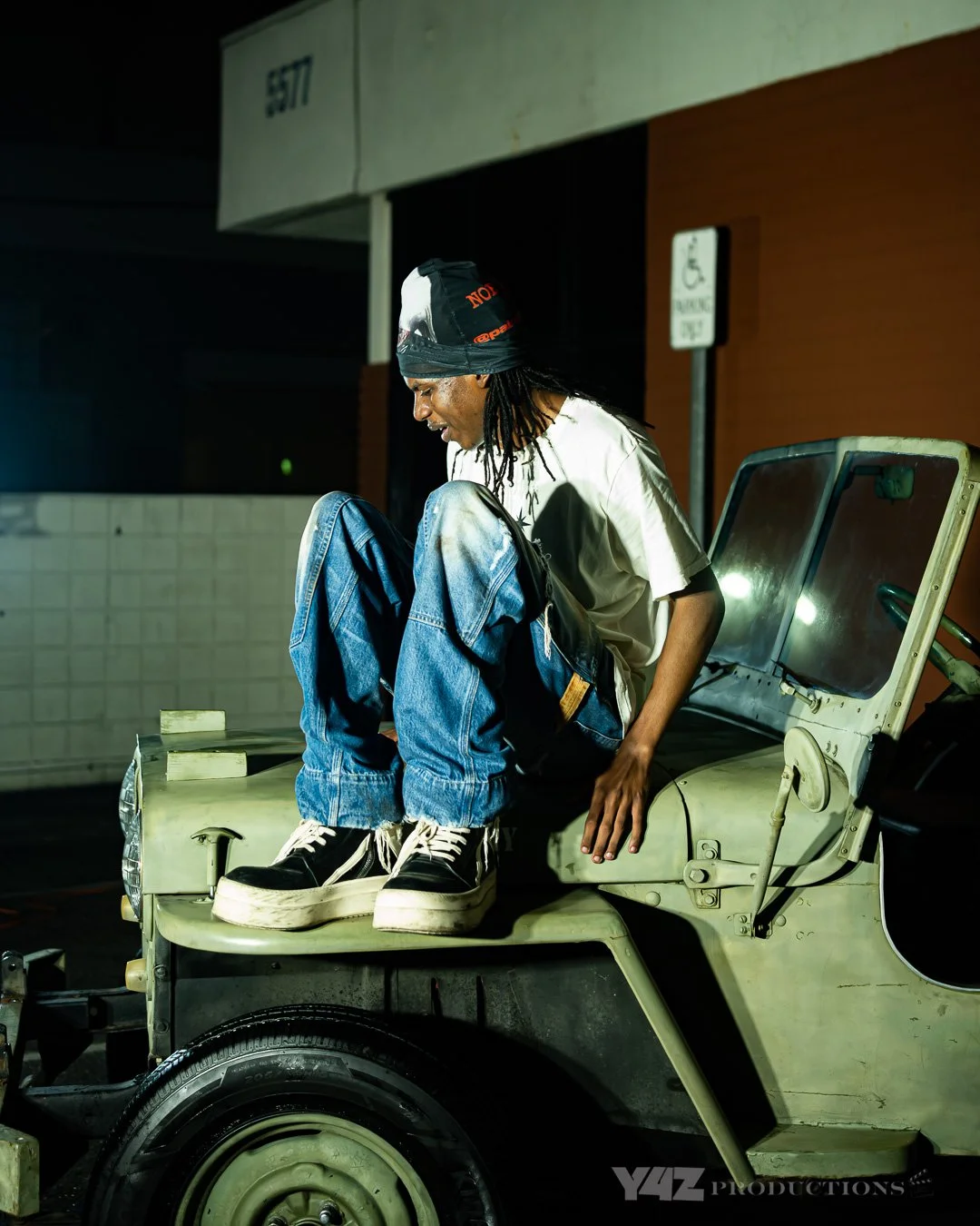 Ezcodylee , wearing a black head wrap, white t-shirt, and jeans, is sitting on the hood of a vintage cream-colored car at night, with a smile on their face.