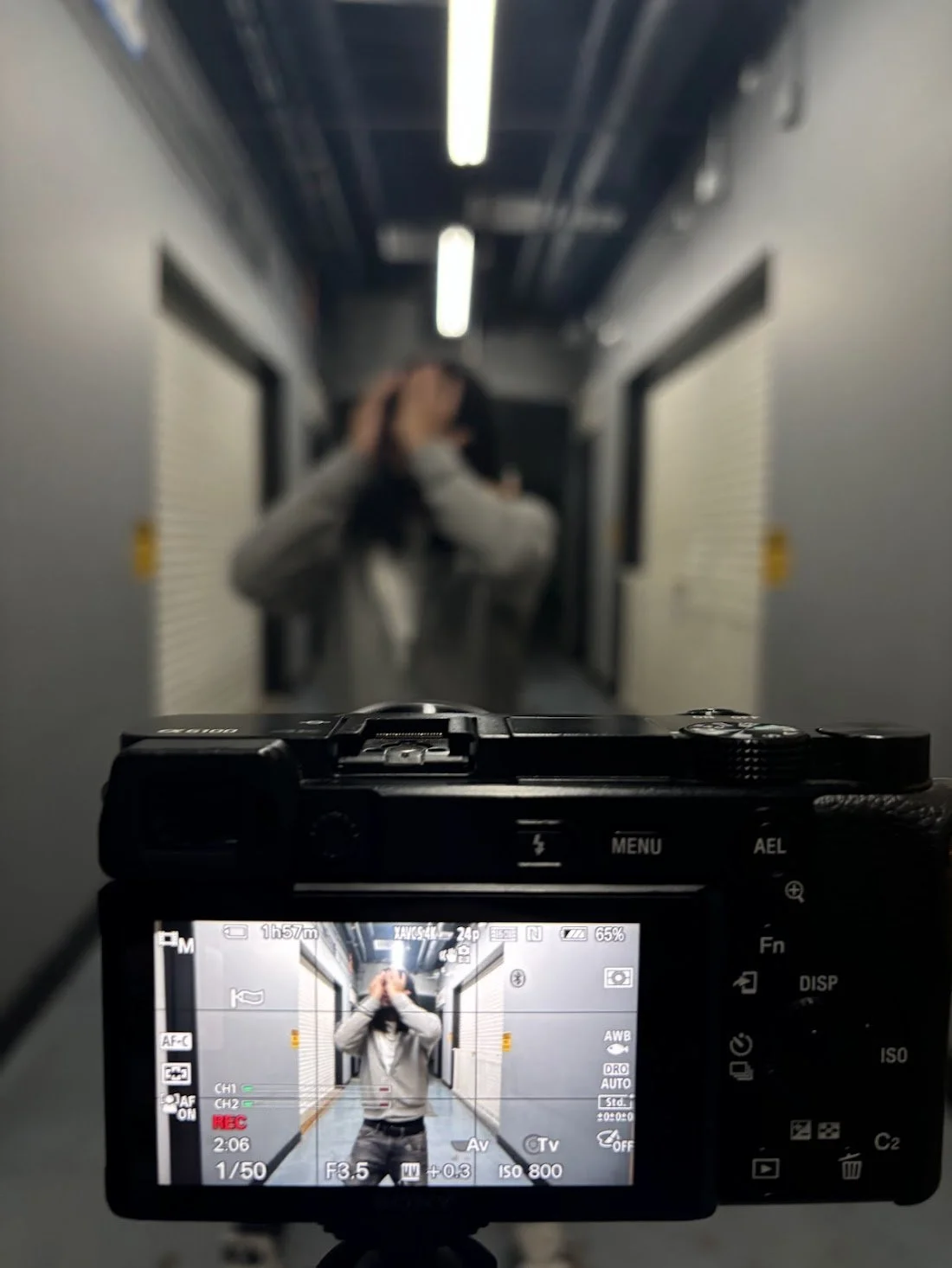 A person standing in a hallway, taking a photo of their reflection in a mirror with a camera. The camera screen showing the person posing with hands near their face.