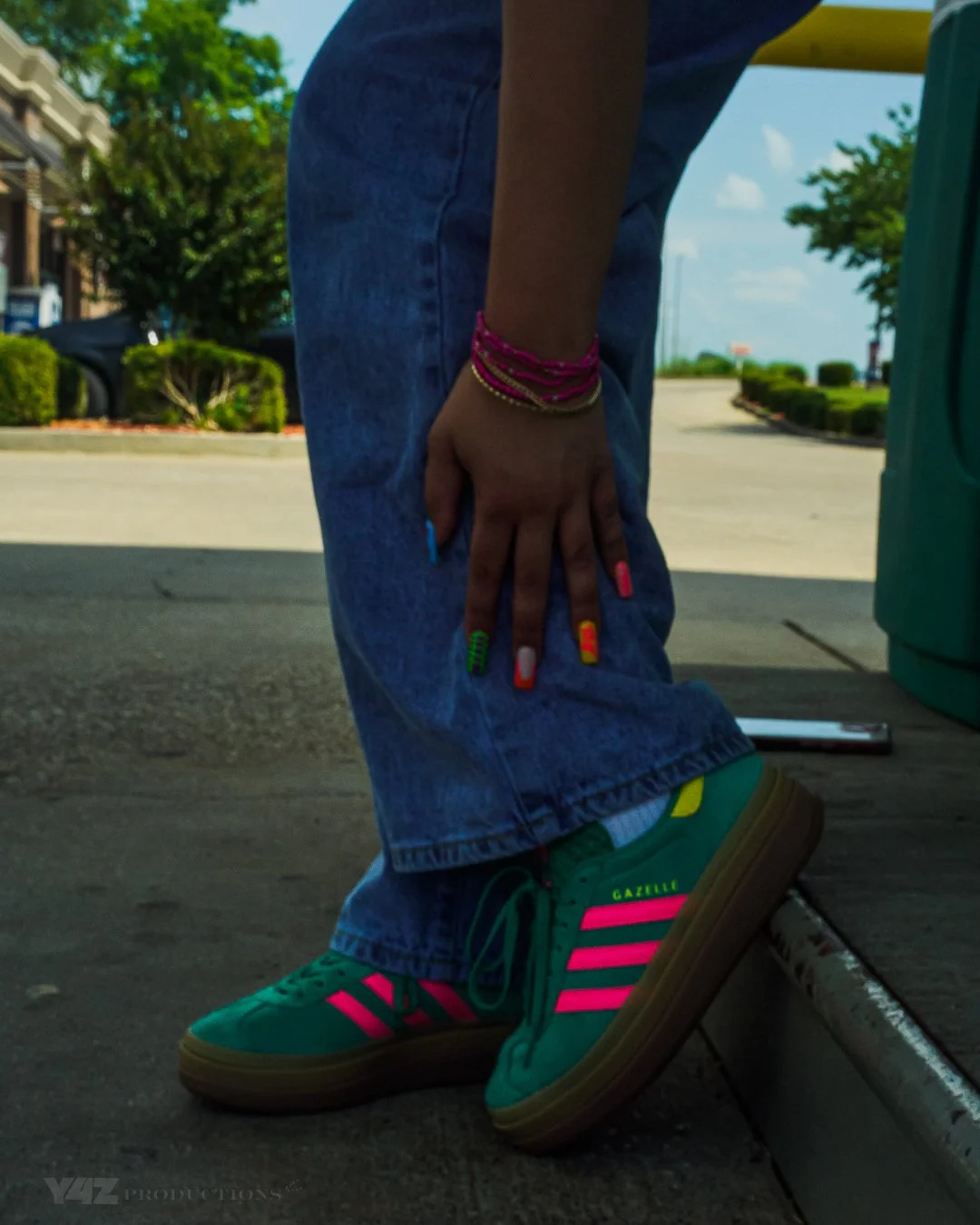 Close-up of a person wearing colorful sneakers, jeans, and wristbands, standing on pavement near a curb with a street scene in the background.
