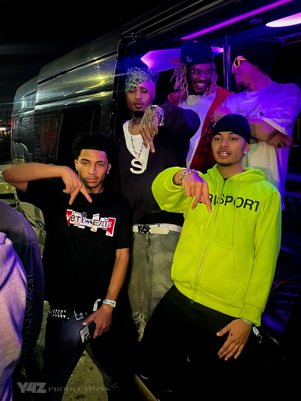 Group of five young men posing at night, standing near a bus with purple and yellow lights, showing casual streetwear and jewelry.