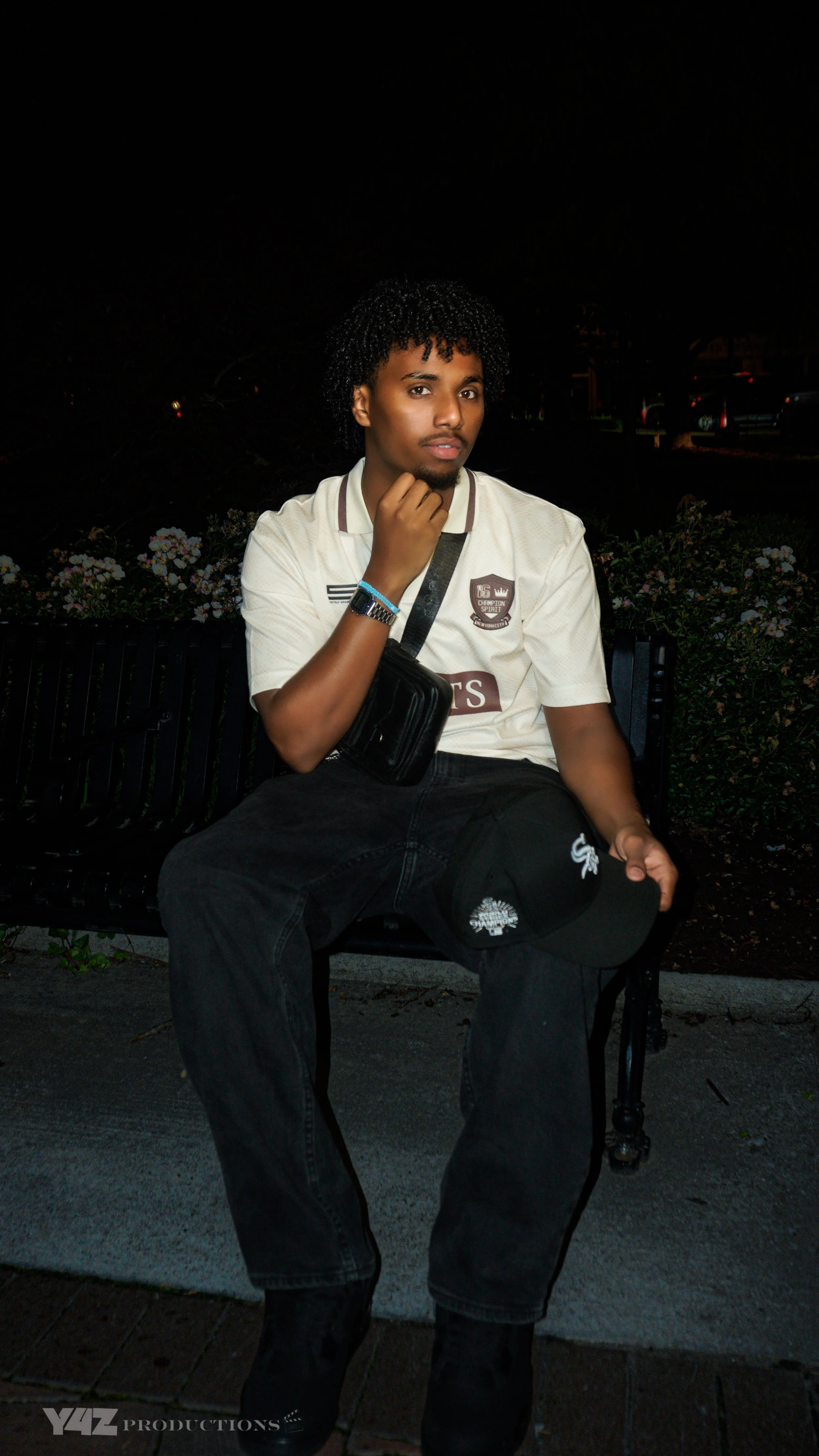 A young man with curly hair and a beard sitting on a black park bench at night, wearing a white polo shirt, black pants, and black shoes, holding a black cap in his left hand and a black shoulder bag across his chest.