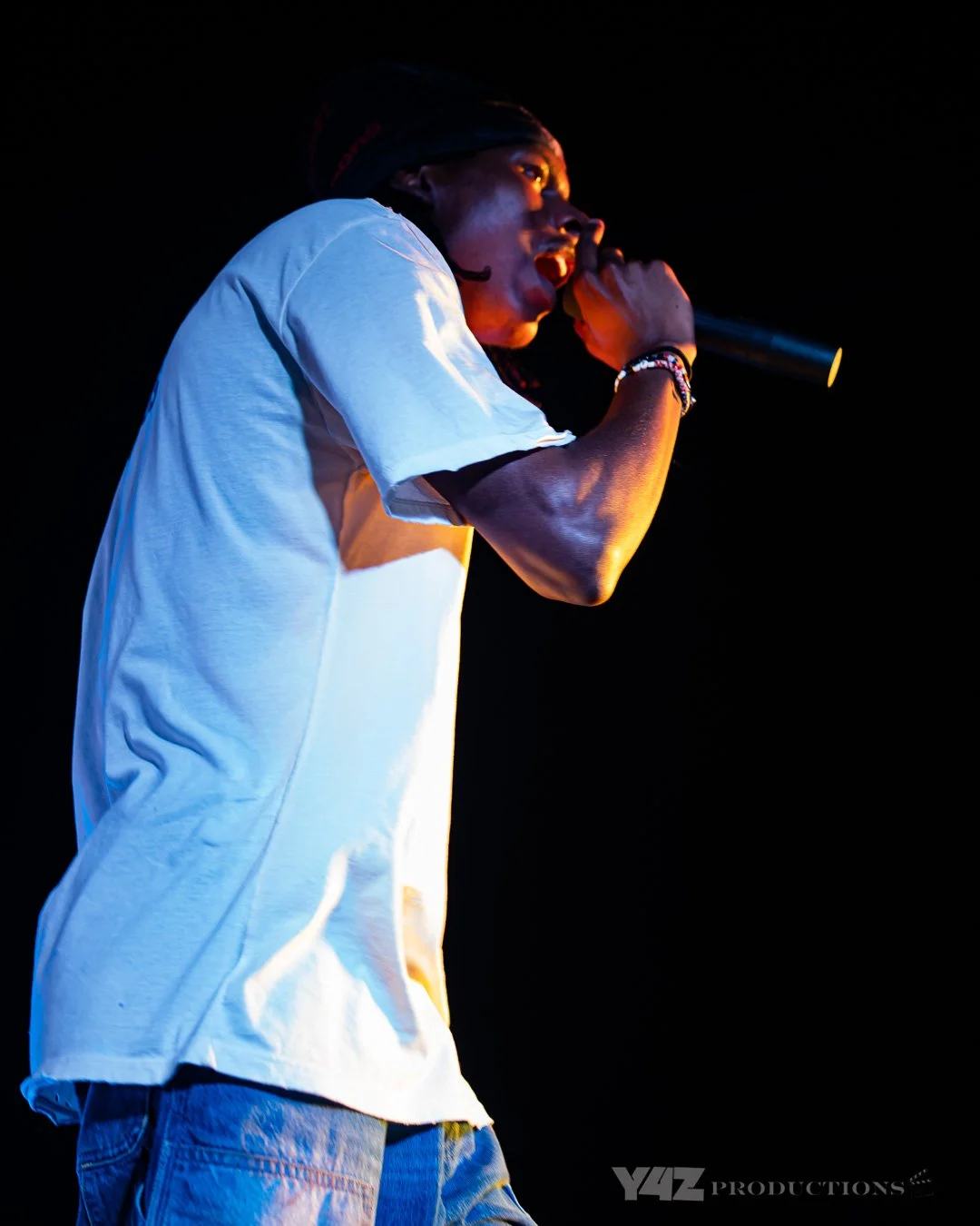 Ezcodylee wearing a white t-shirt and jeans, singing into a microphone on a dark stage.