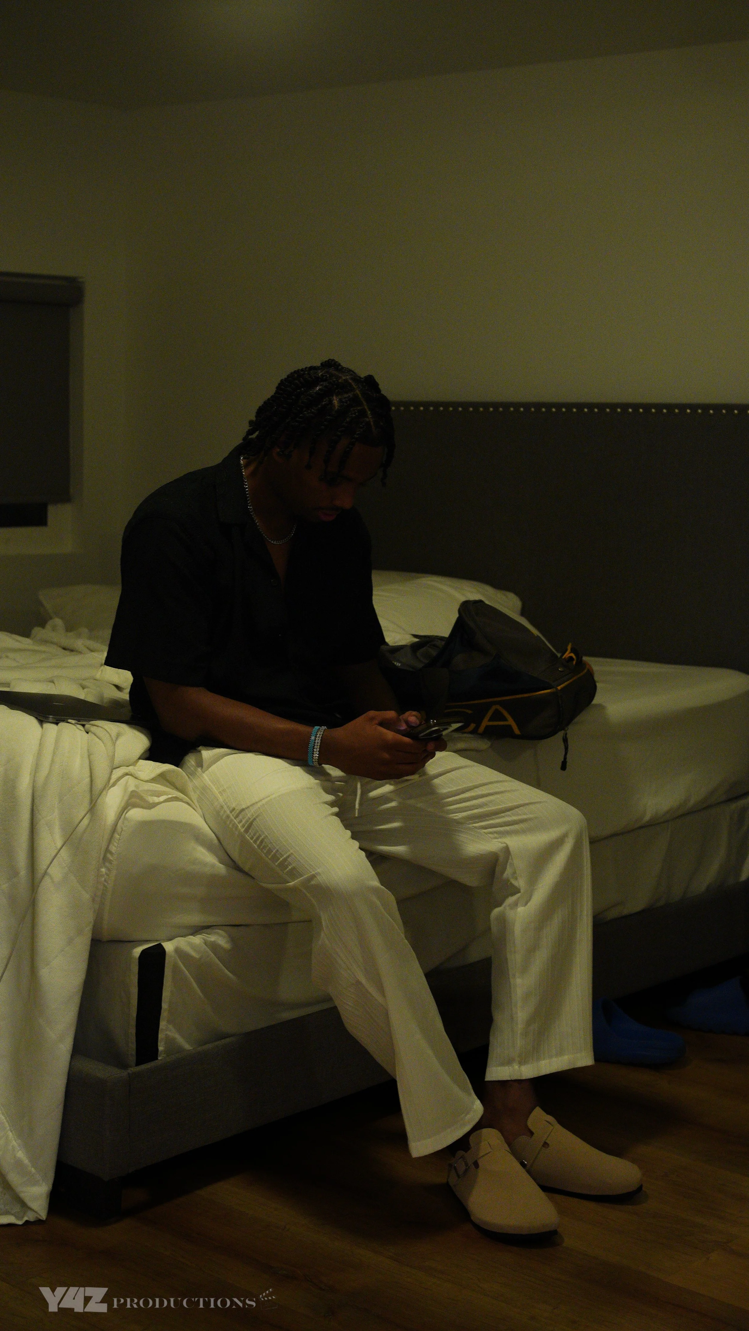 A person sitting on a bed in a dimly lit room, looking at a mobile phone, with a dark backpack beside them and a pair of beige slippers on the floor.
