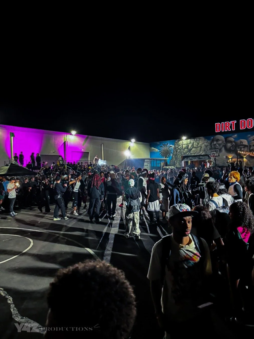 Crowd of people at an outdoor event or concert at night, with colorful lighting and a mural of faces and palm trees on the building wall.