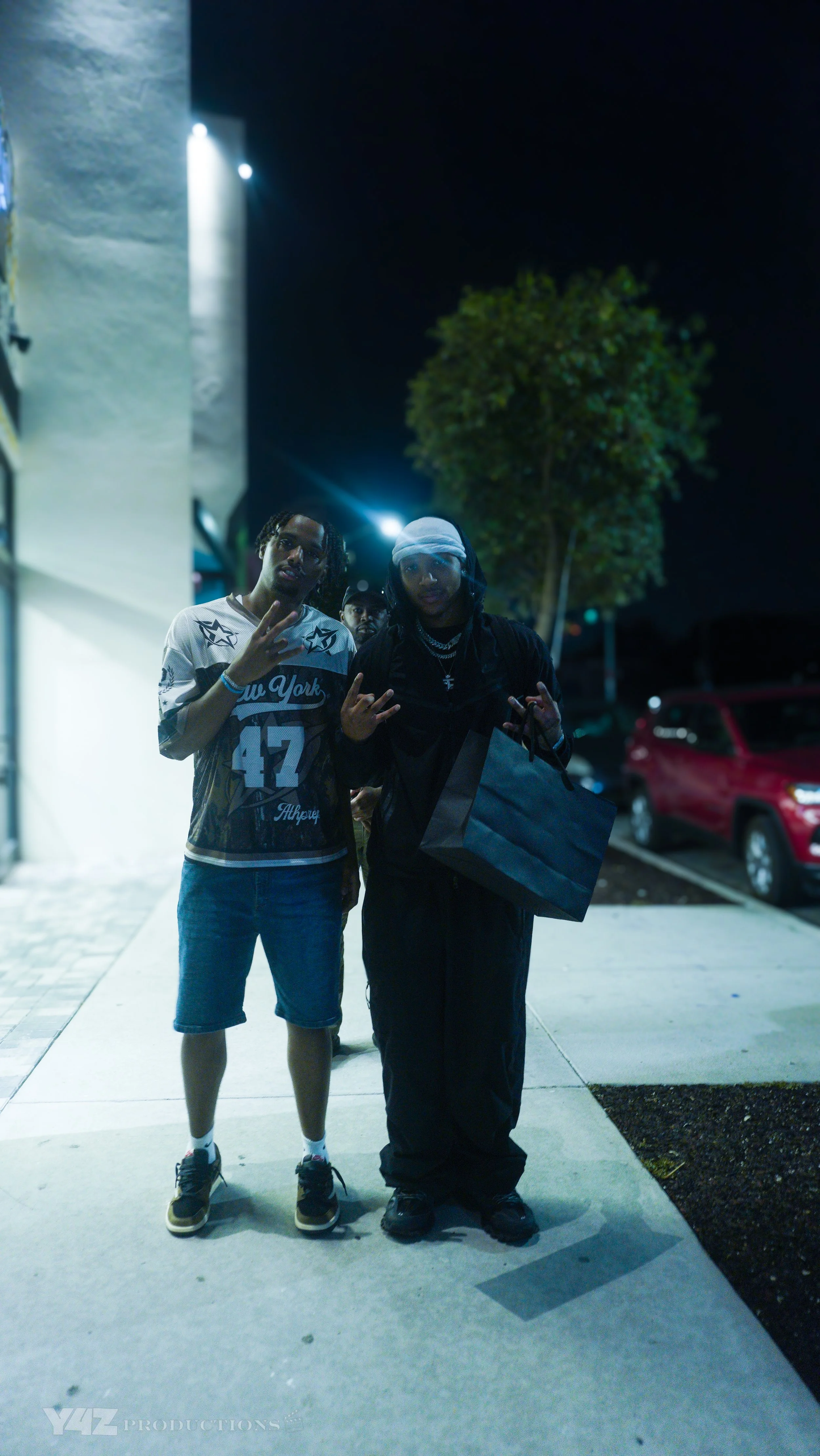Faze Silky standing outdoors at night, one making peace signs with his hand, while the other holds a shopping bag. They are near a parking lot with cars and a tree, illuminated by streetlights.