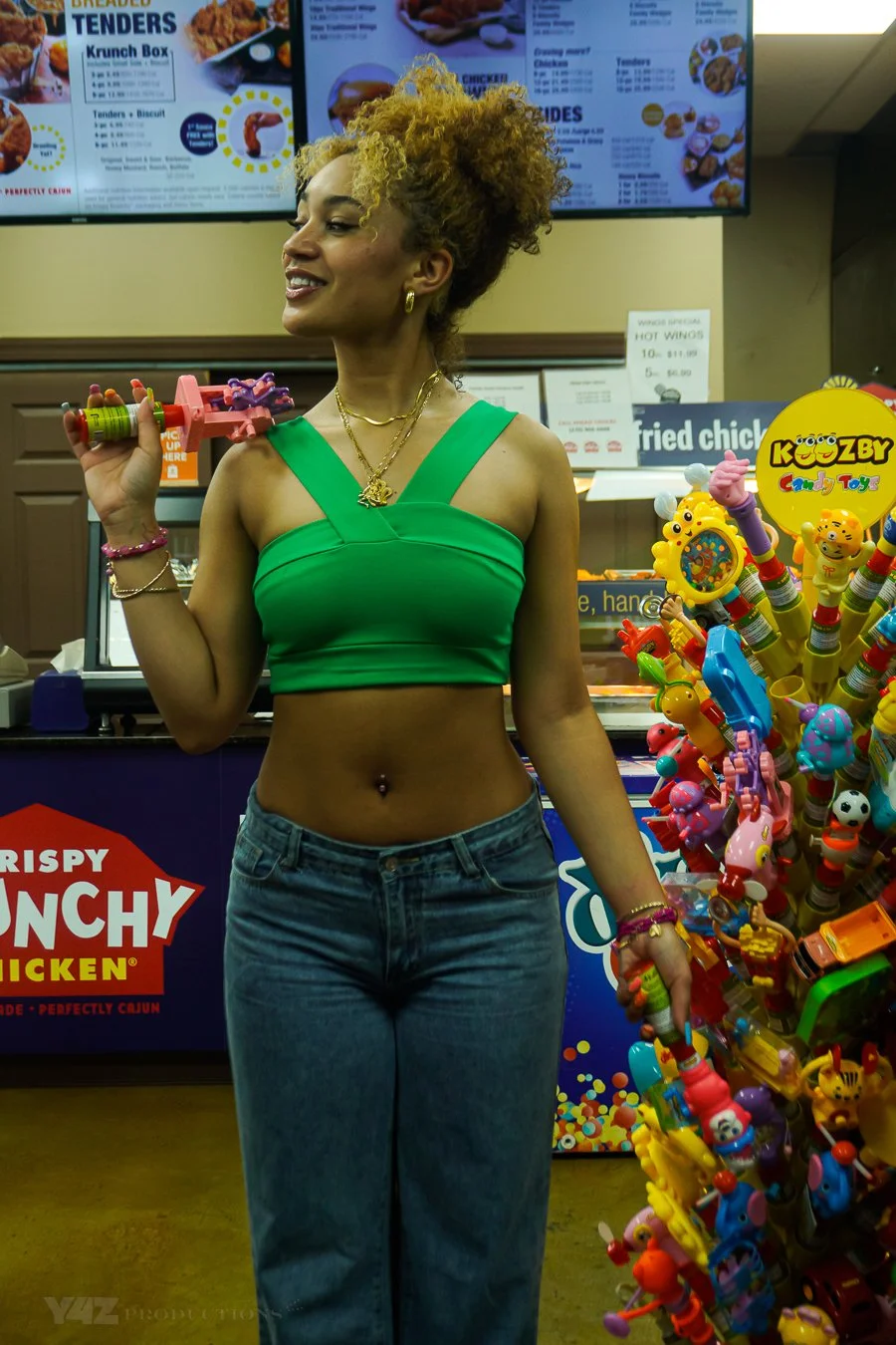 A woman with curly blonde hair wearing a green crop top and blue jeans standing inside a colorful candy store, holding a pink toy gun, with a background of vending machines and a display of various toys and candies.