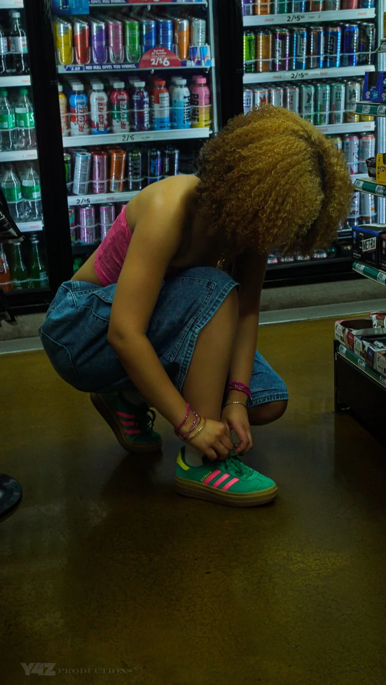 A young woman with curly hair is crouching down in a store aisle to tie her shoelaces. She is wearing a pink top and denim shorts, with colorful sneakers. Behind her are shelves stocked with various canned and bottled beverages.
